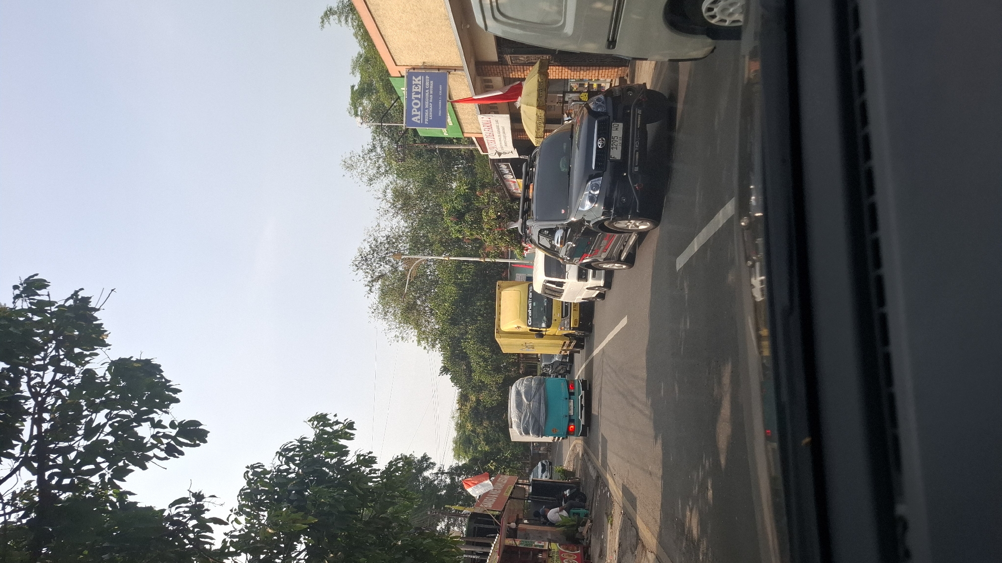The image captures a street scene in Sukasari, Indonesia, on a bright, clear day with a bright sky. The perspective is from inside a vehicle, looking forward through the windshield, which is partially visible along the bottom of the frame. The road ahead is lined with trees on both sides, with some buildings and shops visible on the right.
Traffic is present on the road. In the foreground, to the right, is the side of a light-colored vehicle. Further ahead, in the right lane, is a black SUV with roof racks. Behind the SUV is a white van, and then a yellow truck with "GrahaTrans" written on its side. In the left lane, a turquoise truck with a covered cargo area is driving, and behind it, a small white car is also visible. On the left side of the road, there are some stalls or shops with people sitting and standing. One stall has a red and white flag displayed. A sign for "APOTEK" (Pharmacy) is visible on a building on the right side, with "PRIMA MEDIKA GRUP LENGKAP DAN MURAH" written below it. Another sign below that reads "APOTEK DEA FARMA 2". The overall impression is a busy but not heavily congested street in a commercial or mixed-use area.