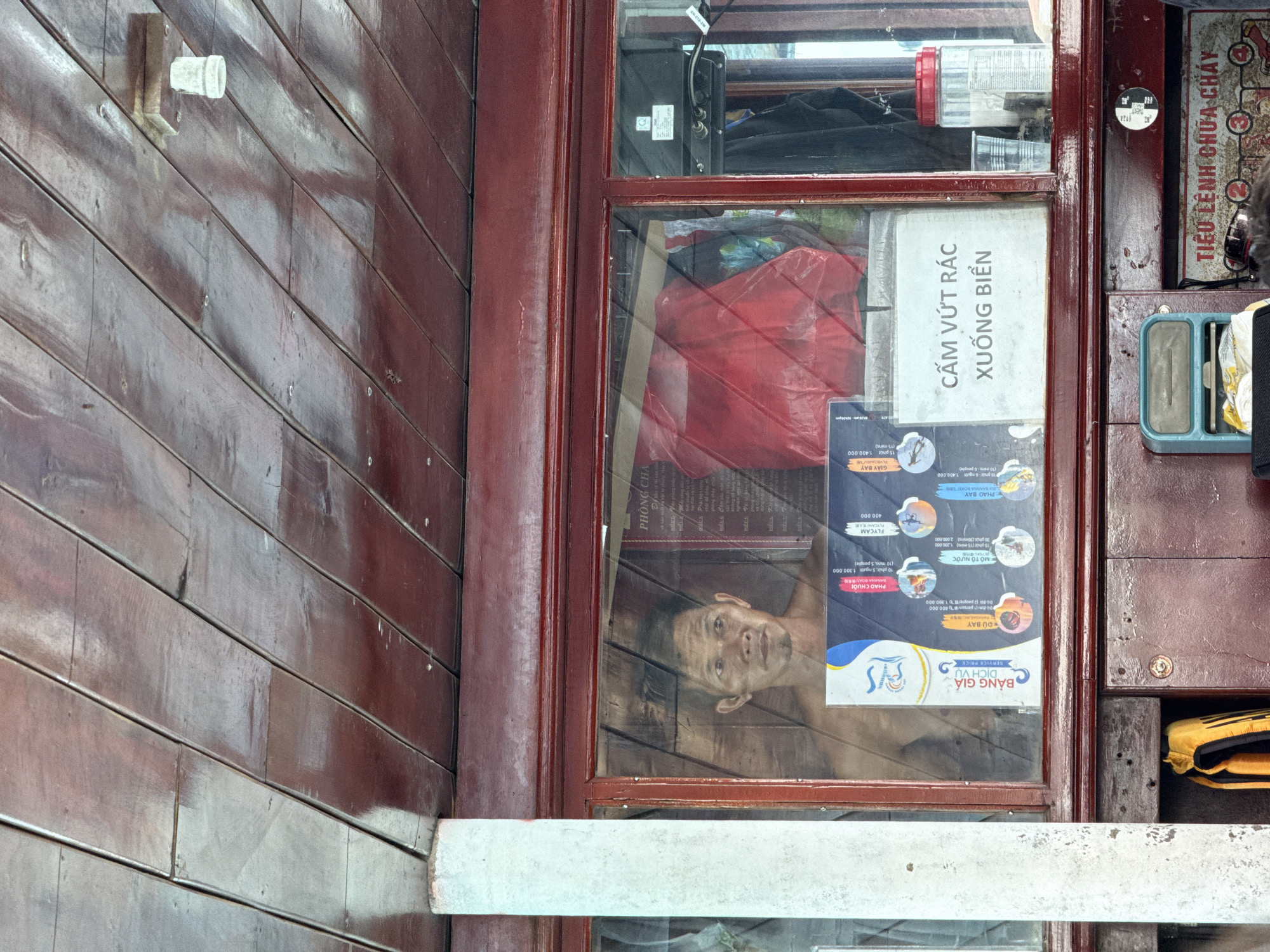 The image shows a storefront window in Bac Nha Trang Ward, Vietnam. The window is wood-framed and reflects the face of a man, possibly the shopkeeper, inside. The window also displays a sign in Vietnamese prohibiting littering on the beach ("Cấm vứt rác xuống biển"). A second sign lists prices for tourist activities like parasailing, jet skis, and banana boat rides. The exterior wall is made of horizontal dark wood planks. A white support beam runs horizontally below the window. Other items visible through the window include a red bag, electronic equipment, and various containers. A sign describing fire safety is also displayed in the storefront window. A sign stating “TIEU LE NH CHUA CHAY” in Vietnamese is also present.