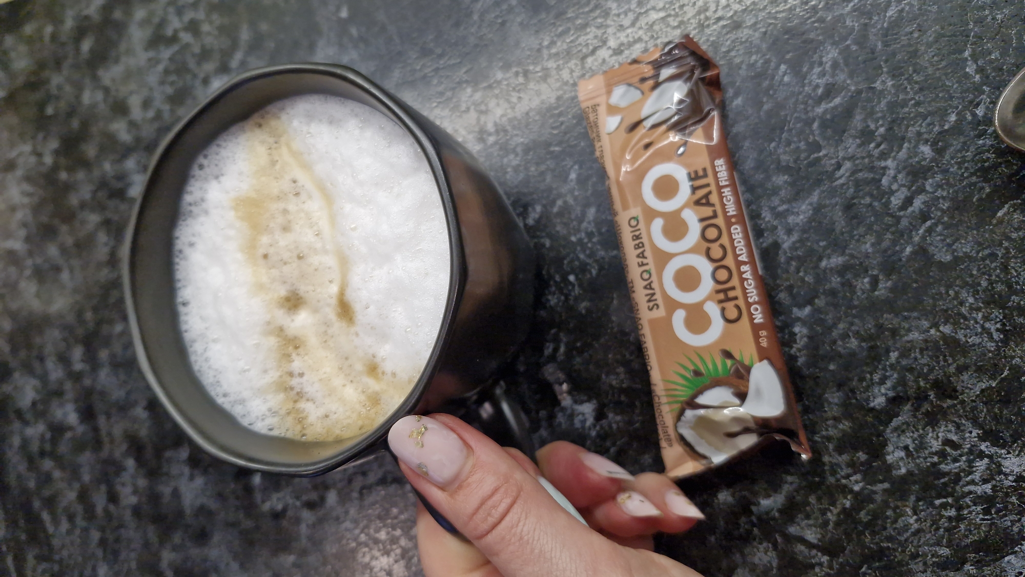 The image depicts a black mug filled with a foamy beverage, likely coffee with milk, placed next to a "COCO Chocolate" bar by "SNAQ FABRIQ." The bar's packaging indicates it contains no sugar and is high in fiber. A person's hand, with painted nails, is holding the mug. The background is a mottled dark gray surface. No information indicates that this image was taken in Otchor, Russia.