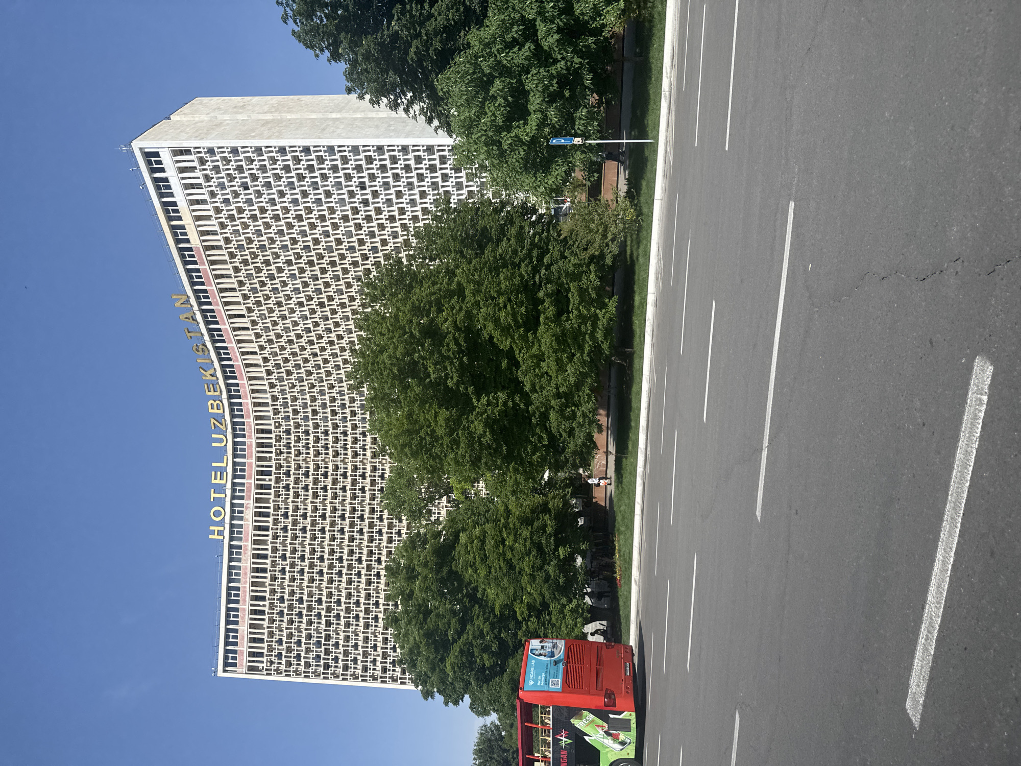 The image depicts a street scene in Kashgar mahalla, Uzbekistan, dominated by the tall, rectangular Hotel Uzbekistan building. The building's facade features a grid-like pattern and the hotel's name is vertically displayed on its side in yellow lettering. Adjacent to the hotel, lush green trees provide a contrast. A road runs alongside the hotel and trees, with clearly marked white lane dividers. A red and green tour bus is positioned on the roadside. The sky is clear and blue, suggesting a sunny day. No people are readily visible in the image.