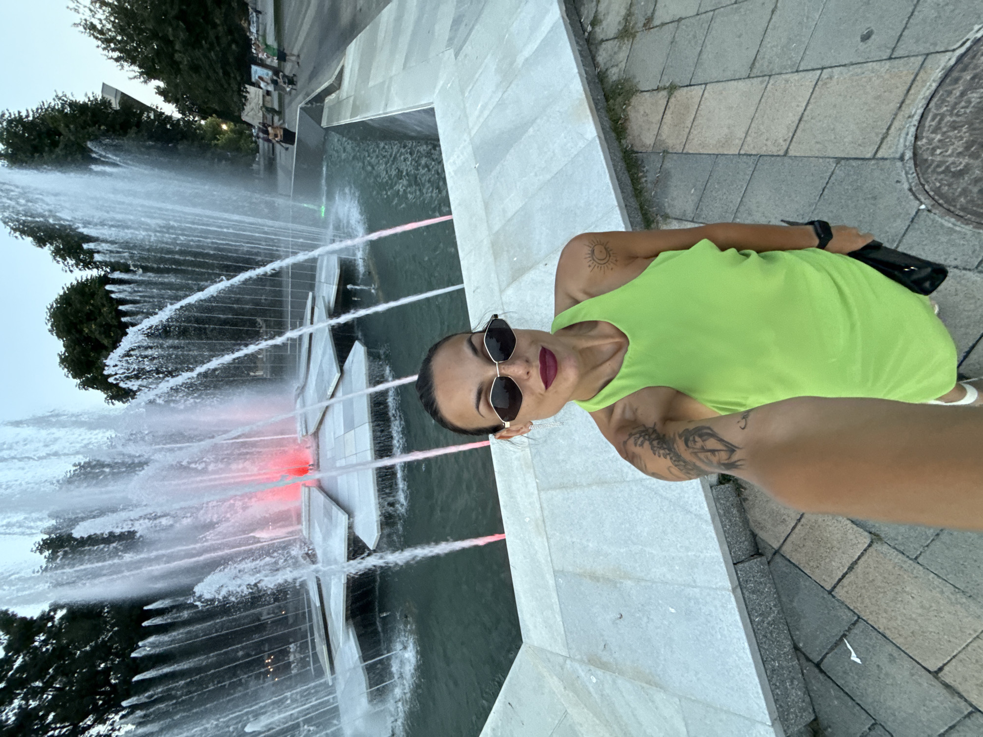 A woman takes a selfie in front of a large, multi-tiered fountain with illuminated water jets in Pleven, Bulgaria. She is wearing a bright green sleeveless dress, sunglasses, and has tattoos on her arm and shoulder. The fountain has a complex design with water cascading over sculpted white stone elements. In the background, trees and a few people are visible. The ground around the fountain is paved with stone tiles.