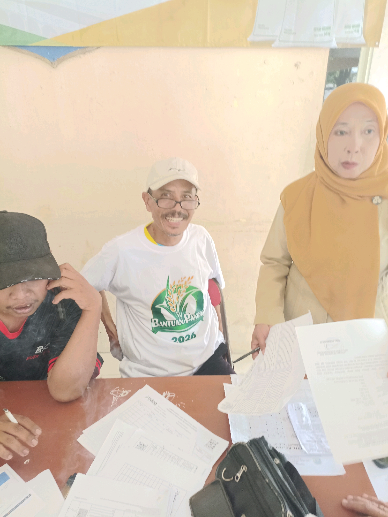 In Guminingrejo, Indonesia, three individuals are gathered around a brown table, interacting with documents. The person on the left, wearing a black baseball cap and a black and red t-shirt, holds a phone to their ear. In the center, a man in a white t-shirt with a "BANTUAN PANGAN 2026" graphic and a white baseball cap smiles at the camera, leaning on a crutch. To the right, a woman wearing a mustard-colored hijab and a beige uniform looks forward. Scattered across the table are various documents, a black bag, and a pen, suggesting an administrative or registration process. The background features a light-colored wall with a colorful abstract mural at the top.