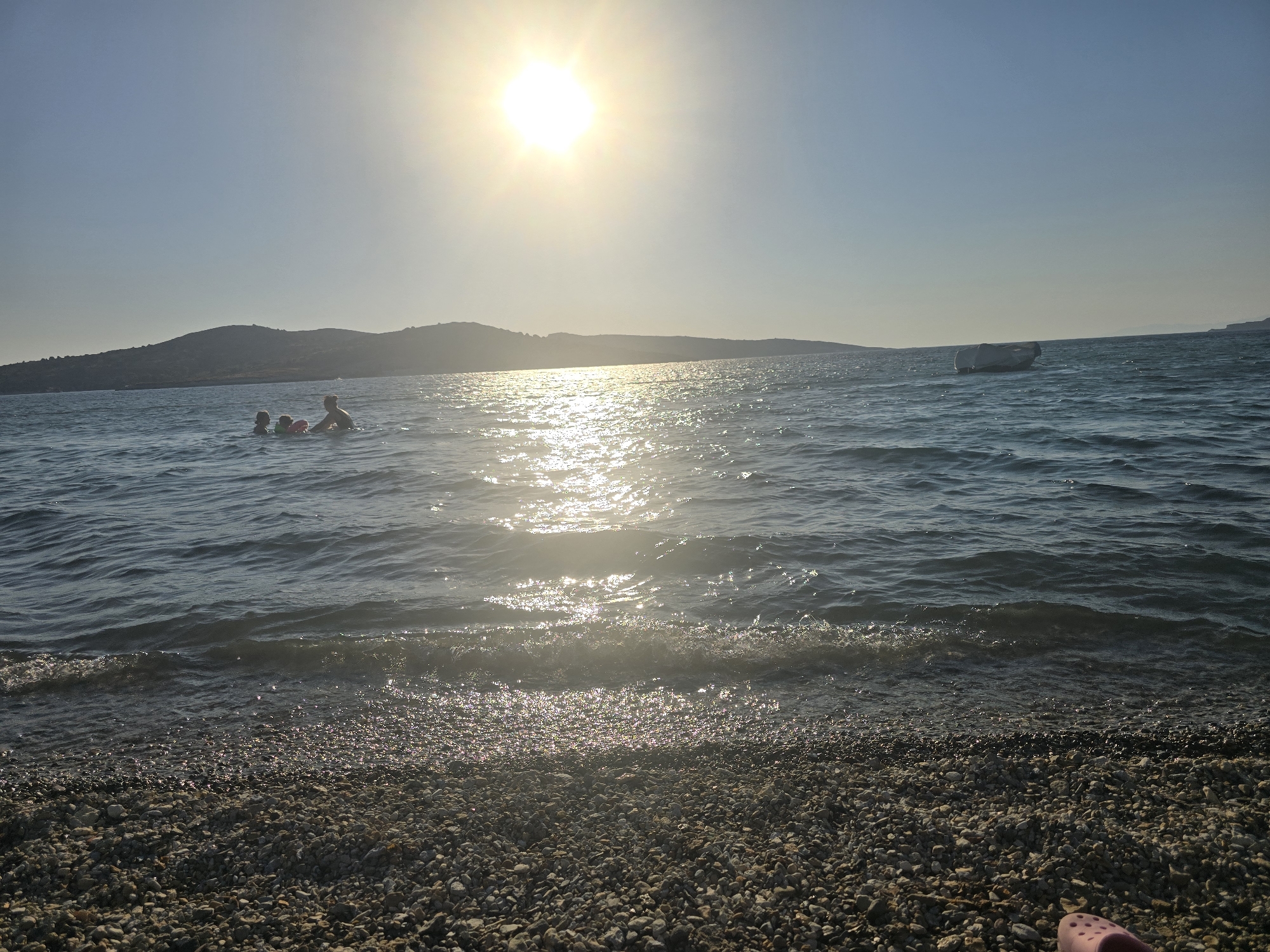 A sunny, wide shot shows a pebble beach in the foreground, with the Aegean Sea stretching out beyond. In the middle distance, three people are in the water, one appearing to be an adult holding onto a child with a pink inflatable ring. A white, possibly inflatable, object or small boat is visible further out in the sea. In the background, a low, hilly coastline is silhouetted against a clear, pale blue sky. The sun is high and bright, casting a strong glare on the water. The scene evokes a peaceful, late afternoon summer day at a coastal location in Foça, Turkey.