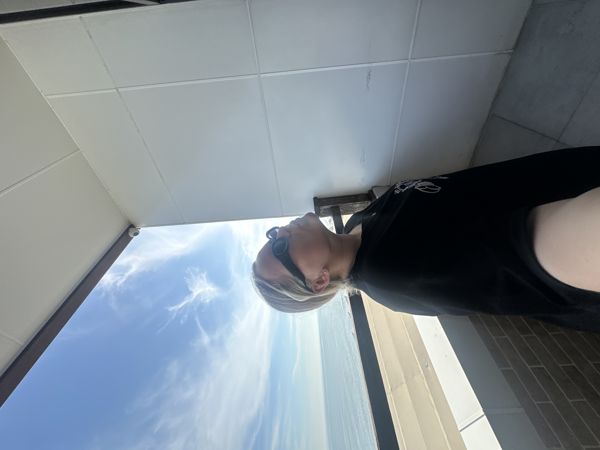 A woman with blonde hair, wearing black sunglasses and a black top, stands on a modern balcony, captured in a selfie perspective with her lower arm visible. Her head is tilted back, and her gaze is directed upwards and to the right, towards a bright blue sky featuring wispy white clouds and a prominent sun glare in the upper right. The balcony overlooks the Black Sea, consistent with its location as a coastal property in Lermontovo, Russia. The balcony structure includes a dark horizontal top railing, a patterned lower railing resembling brickwork, and white paneled walls with minor scuff marks. A dark wooden support post is visible behind the woman. The ceiling of the balcony overhang is white and paneled, with a small, white, dome-shaped security camera mounted near the corner. A grey tiled floor is visible at the bottom right of the frame. The sea extends to the horizon under a clear sky.