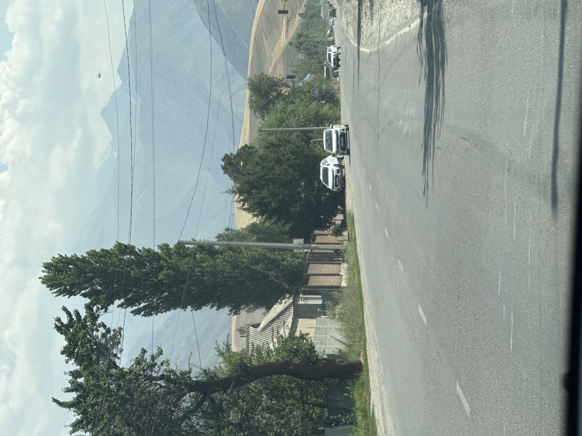 In Koi-Tash, Kyrgyzstan, a two-lane asphalt road stretches into the distance, flanked by sparse vegetation and buildings on the left. The road is marked with white dashed lines, indicating lanes of traffic. Several cars, including a white SUV and a white sedan, are visible on the road. In the background, rolling hills and majestic mountains dominate the landscape, with power lines crisscrossing the sky. A small, single-story brick house with a grey tiled roof is situated on the left side of the road, behind a fence. The sky is partly cloudy.