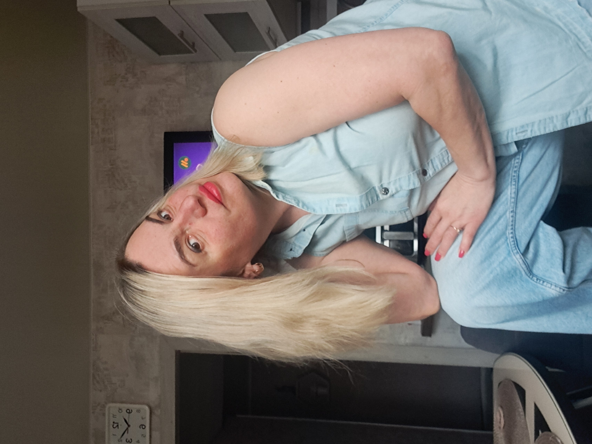 A woman with blonde hair and red lipstick is wearing a light blue denim shirt and jeans. She has a ring on her finger and her nails are painted red. She is in a room with a clock on the wall that reads 1:57. There are cabinets in the background and a television. The location is unknown. The city of Elektrostal, Russia is not discernible from the image.