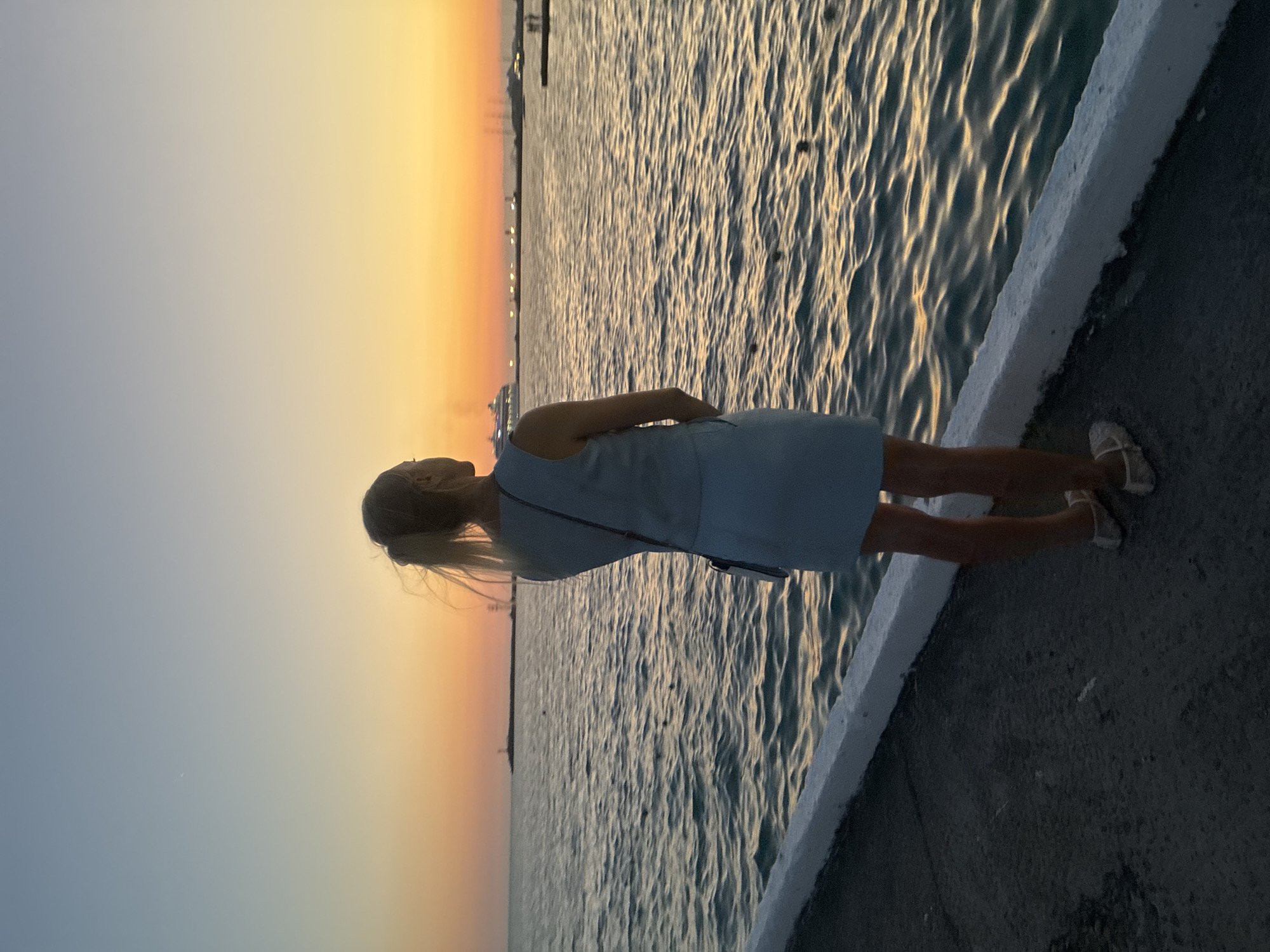 A full shot captures a woman standing on a pier, facing away from the camera, looking out at the sea during sunset. The sky transitions from a pale blue at the top to a vibrant orange and yellow hue near the horizon, indicative of a sunset over the sea. The water is calm with small ripples, reflecting the colors of the sky. In the distance, a large cruise ship is visible on the horizon, along with a lighthouse and some structures that suggest a port or coastal city. The woman is wearing a light blue dress and a small crossbody bag. She has blonde hair tied back in a ponytail. The pier is made of dark asphalt with a white concrete edge. The scene suggests a peaceful moment of contemplation at the end of the day, possibly in the city of Sochi, Russia, given the presence of a cruise ship and coastal landscape often seen in that region.