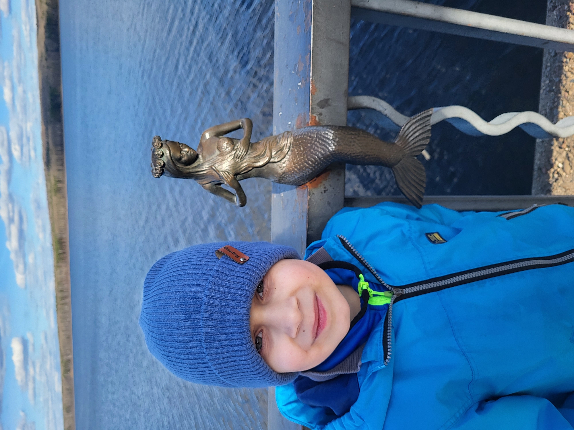 A boy wearing a blue hat and a blue jacket is next to a bronze mermaid statue on a bridge railing. The background shows a body of water (possibly a river or lake) and some trees in the distance. The photograph was taken near the city of Chaikovsky, Russia, where a similar mermaid statue is known to exist along the river.