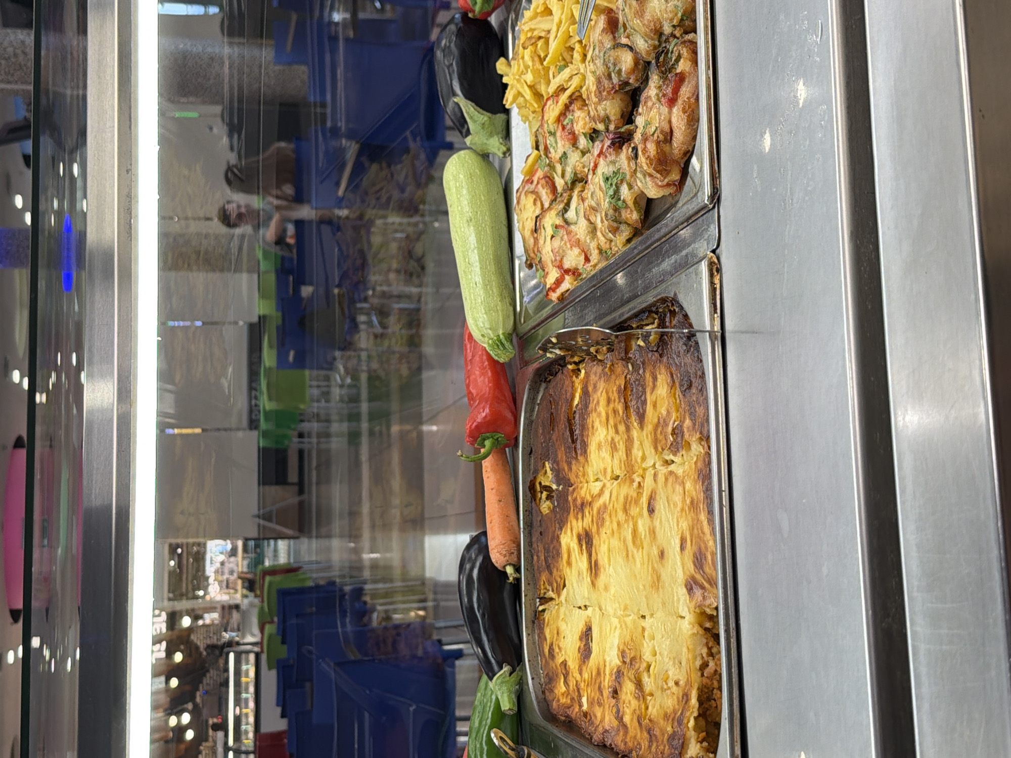 The scene depicts a self-service hot food counter in a cafeteria located in Plovdiv, Bulgaria. In the foreground, two large stainless steel trays present hot dishes. The left tray contains a baked casserole, possibly Moussaka, characterized by a golden-brown top crust, with a serving utensil on its left. The right tray holds multiple individual portions of a baked item topped with cheese and herbs, alongside a mound of golden french fries; a serving fork is partially visible to the right of the fries. Resting on the counter surface between the food trays and the viewer are a green zucchini, two dark purple eggplants, an orange carrot, and a red bell pepper. The counter surface below the trays is stainless steel.
Through the clear glass barrier and its reflections, a dining area is visible in the midground, furnished with multiple blue chairs and tables, with some green chairs further back. Two individuals are discernible in the mid-right through the glass: one male facing left and a female with long brown hair facing away from the viewer, appearing to be in conversation or interaction within the dining space. The ceiling, reflected in the upper portion of the glass, shows numerous recessed circular lights and a distinct pink oval fixture. Further into the background, additional counters and architectural elements of the establishment are faintly visible. The overall environment is well-lit.