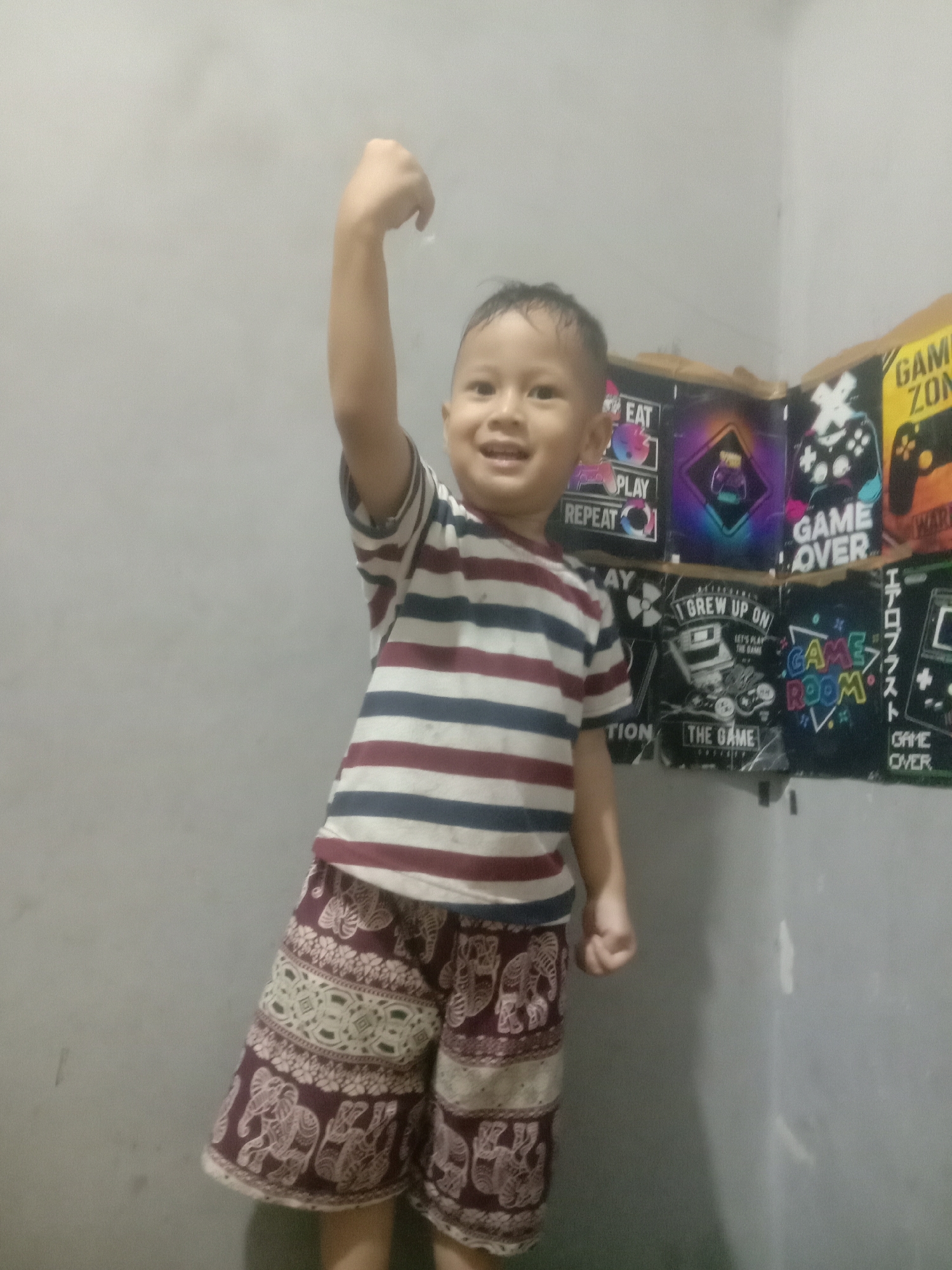 A young boy, possibly Indonesian, stands in a room with gray walls. He is wearing a striped t-shirt with horizontal bands of dark blue, white, and maroon, and patterned shorts featuring elephants and geometric designs in maroon and white. His right arm is raised in a victory pose, and he is smiling. Behind him, a wall is adorned with several posters related to video games, including one with the text "GAME ZONE" and others with imagery of controllers and game-related graphics. The lighting suggests an indoor setting, possibly a bedroom or play area. No specific location within Cianjur, Indonesia can be determined from the image.