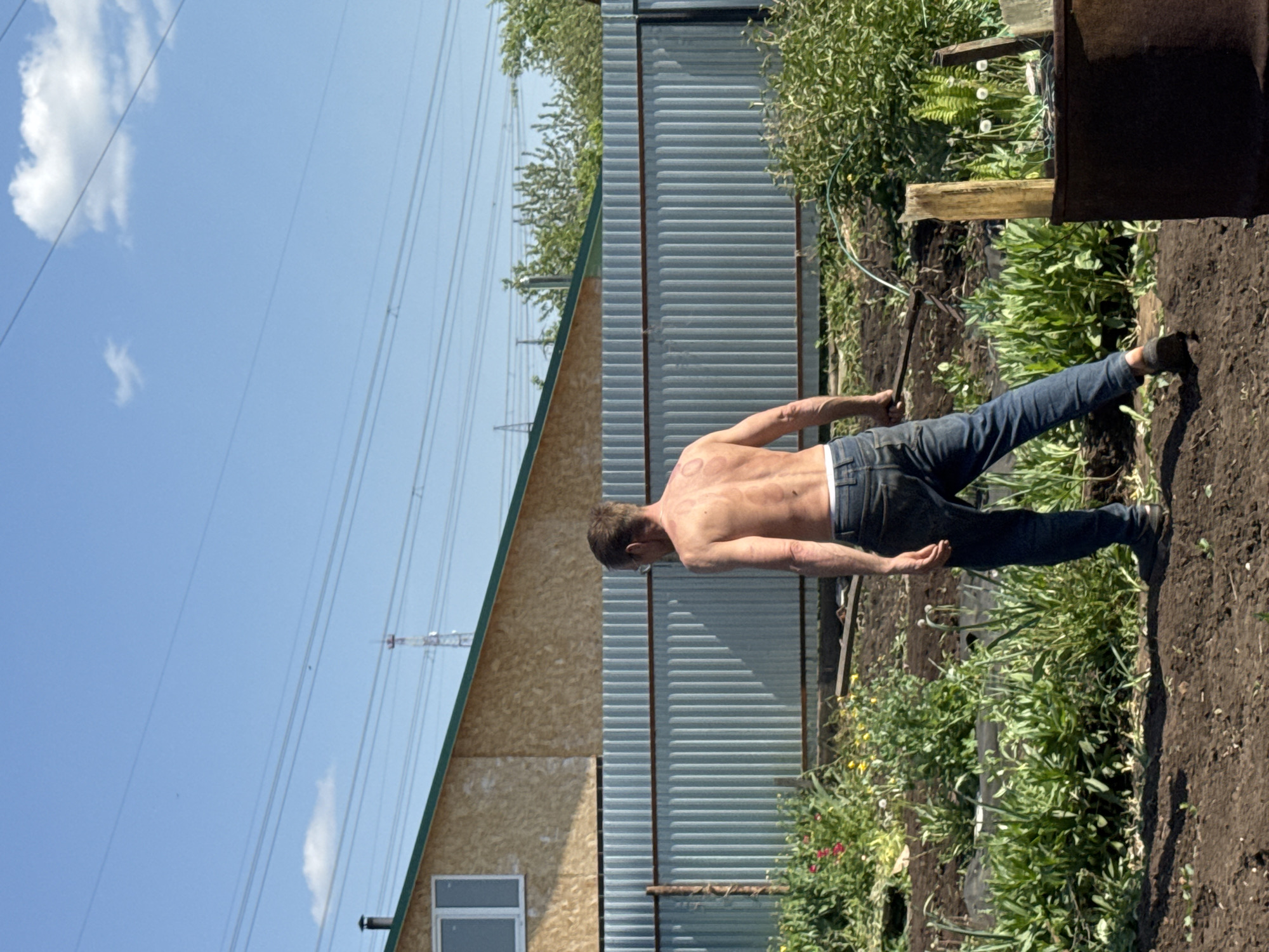 A man is pictured in an outdoor setting near a shed or small building. The man is shirtless, wearing blue jeans and shoes, and is positioned in the right half of the frame. He appears to be working in a garden area, possibly turning over soil with a tool in his hand. Vegetation is visible around the man and the plot he is working on. A building with a corrugated metal exterior is behind him, alongside a roof and an exterior wall made of wood. Overhead power lines are visible against a clear blue sky. Based on the request, this scene is located in Chaikovsky, Russia.
