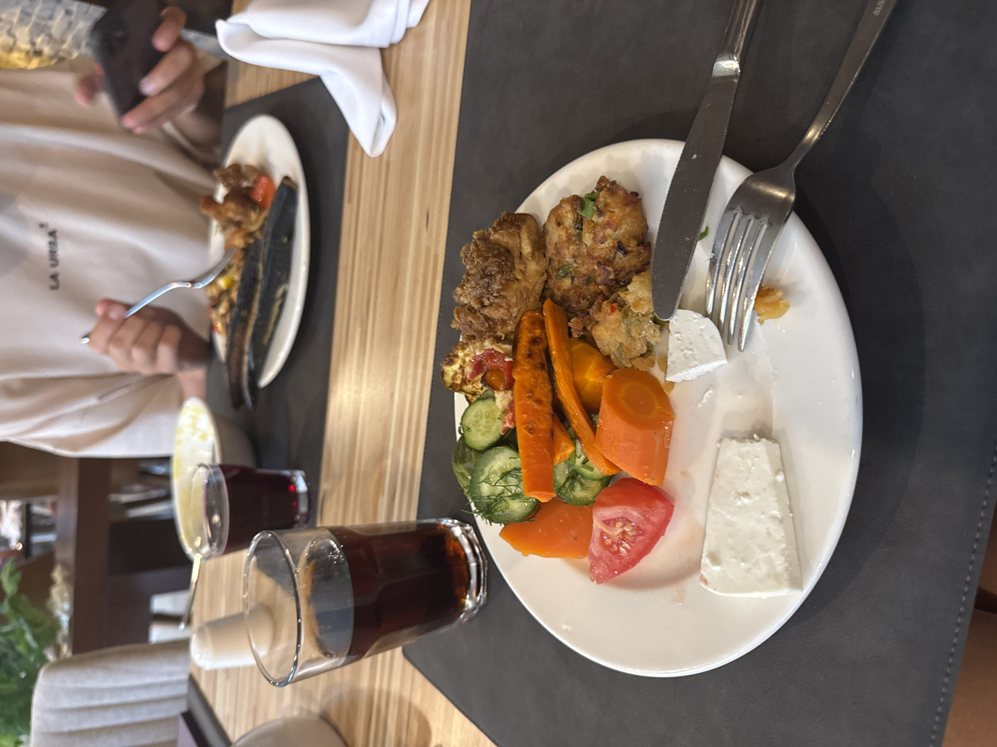 A table setting for a meal is depicted, featuring a white plate filled with food, a glass of dark beverage, and cutlery. The food on the plate consists of sliced carrots, cucumbers, tomatoes, cheese, and fried patties. A person is visible in the background, holding a smartphone and a fork, engaged in eating and using the device. The setting appears to be an indoor dining area, possibly a restaurant or hotel, with a wooden table and chairs. There is no specific information in the image to indicate the location is Aksu, Turkey.