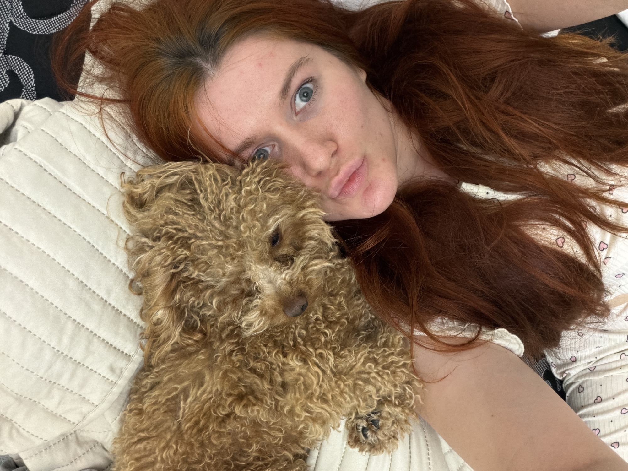 A young woman with red hair and a small, light brown poodle lie on a cream-colored quilted surface. The woman is lying on her back and taking a selfie with her phone, with her head turned towards the camera. Her lips are pursed, and her blue eyes are wide open. The poodle is resting its head on her shoulder, with its face partially obscured by her hair. The woman is wearing a white top with small pink hearts. The scene appears to be indoors, likely a bedroom, given the quilted surface which resembles a bedspread. The lighting is natural and soft, suggesting it may be daytime. There is no visible indication of the location within Krasnodar, Russia.