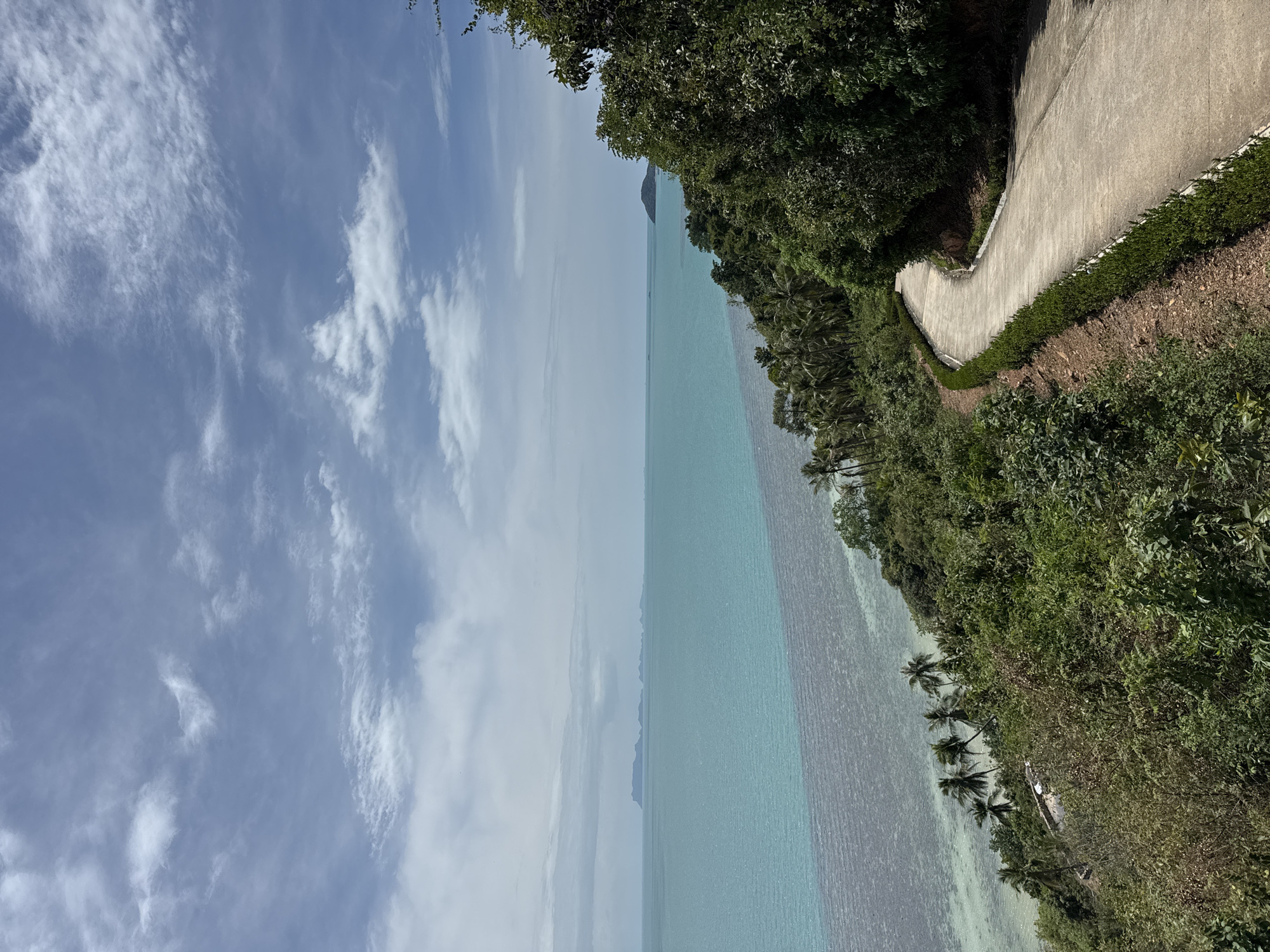 The image captures a view of the ocean and a coastline. The sky, with light white clouds scattered across a blue backdrop, takes up a portion of the left side. A body of water occupies the central area, transitioning from light turquoise to deeper hues, with what appears to be a distant island visible in the far background. The right side depicts a coastline densely covered with greenery, including trees and shrubs. A narrow road runs parallel to the coast, winding through the vegetation. The scene is set in Baan Taling Ngam, Thailand. No people are visible in the frame.
