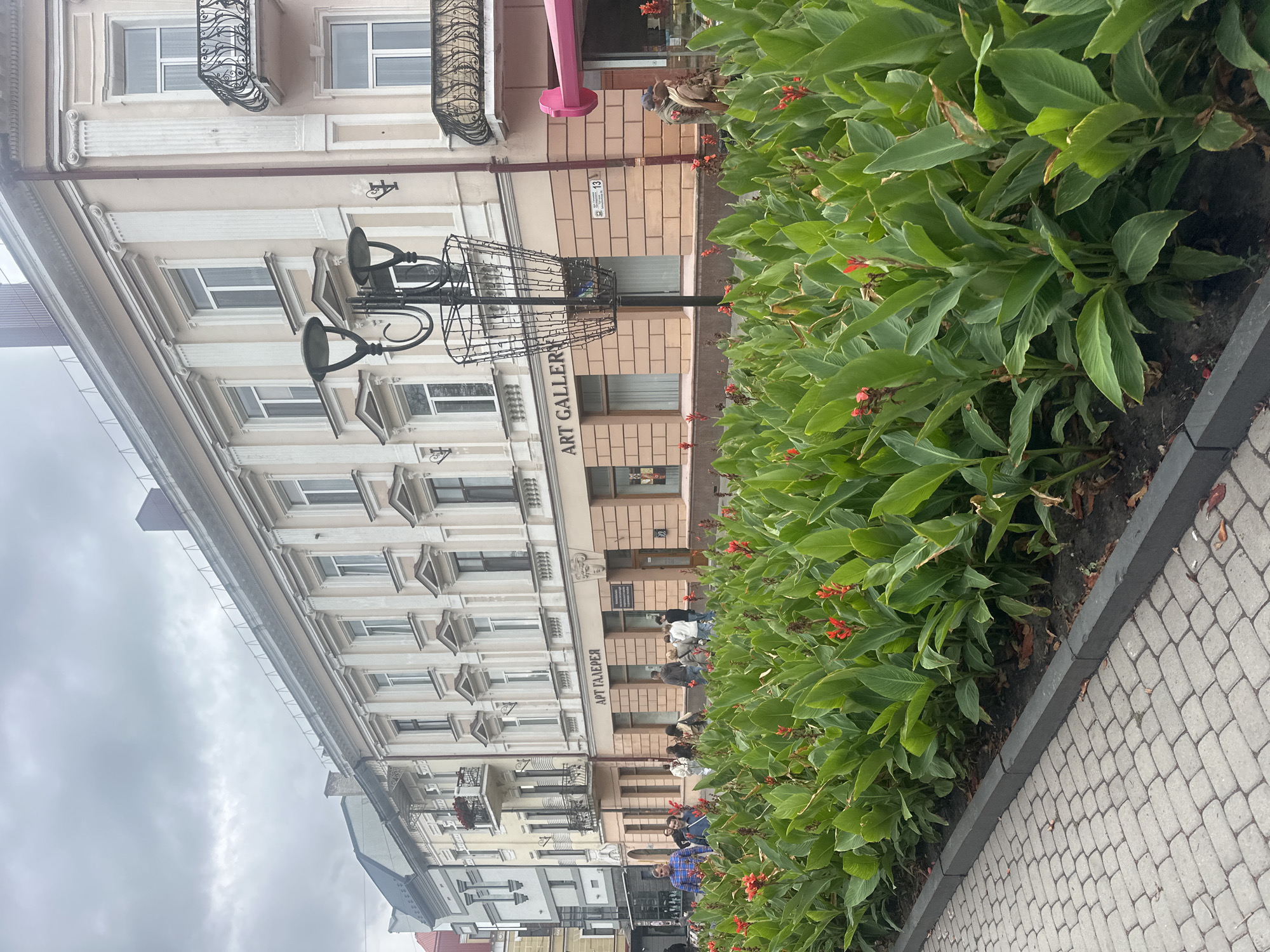 The image captures a street scene in Ternopil, Ukraine, focusing on a row of buildings with an "ART GALLERY" sign. In the foreground, a bed of lush green plants with scattered red flowers lines the sidewalk, which is paved with gray cobblestones. People are visible walking along the sidewalk in front of the buildings, some interacting in small groups. A vintage-style street lamp with a basket-like fixture stands prominently in front of the gallery. The architecture of the buildings suggests an older European style, with ornate details on the facades and balconies. The sky is overcast, indicating a cloudy day. The overall impression is of a pleasant urban environment with a touch of cultural appeal.