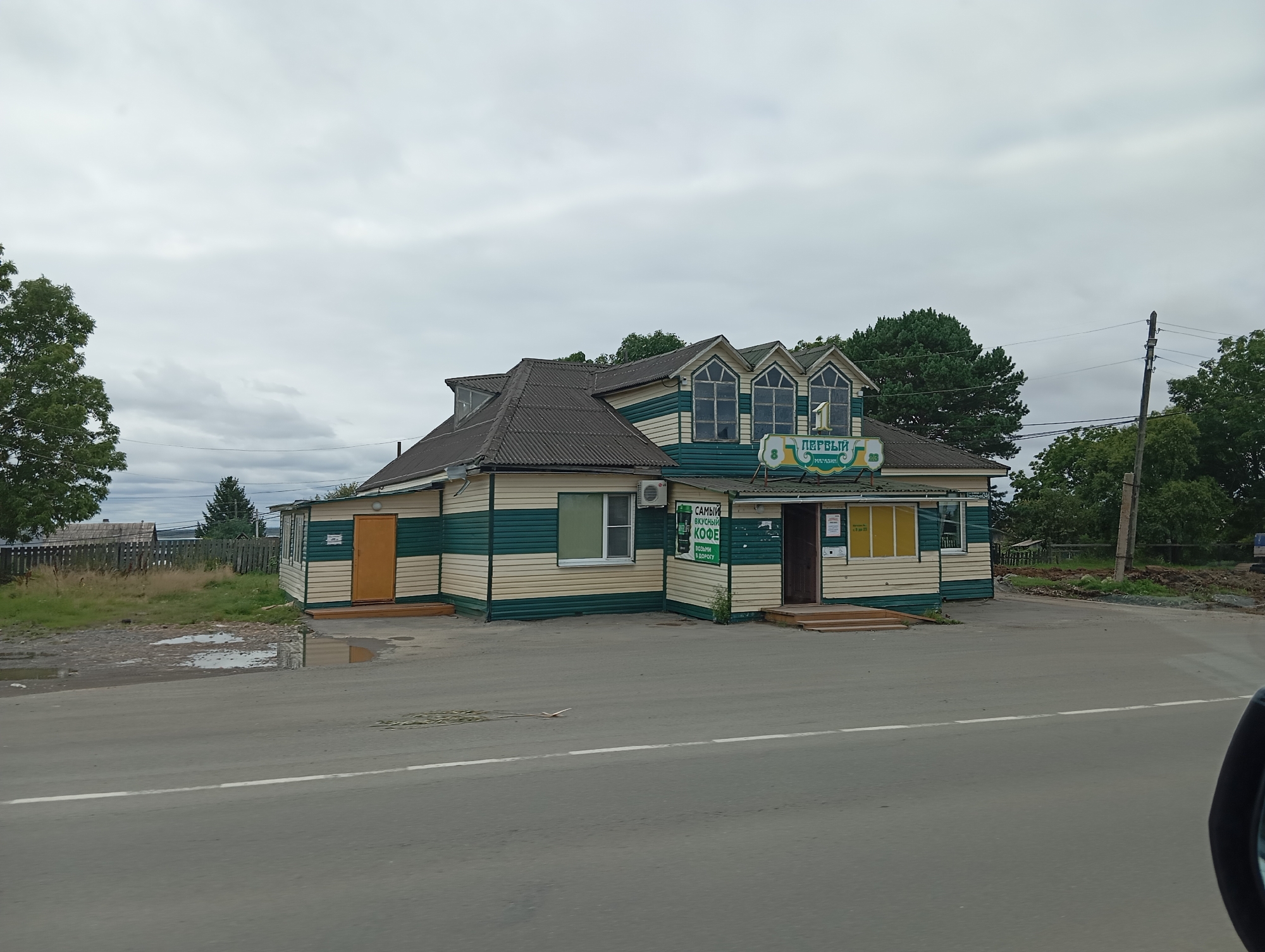 A two-story building, painted with horizontal green and beige stripes, is situated on a paved road in what appears to be a rural or small-town setting. The building has a dark-colored, gabled roof with dormer windows. A sign above the entrance reads "ПЕРВЫЙ МАГАЗИН" (First Store) in green lettering. To the left of the main building, there is a smaller, attached structure with an orange door. A small, single-story building with an orange door is also visible further to the left. The ground in front of the buildings is a mix of grass, gravel, and puddles of water. Trees and a wooden fence are visible in the background. The sky is overcast with gray clouds. The image is taken from a low angle, possibly from a vehicle, as the edge of a car mirror is visible in the lower right corner. The location is identified as Mayak, Russia.