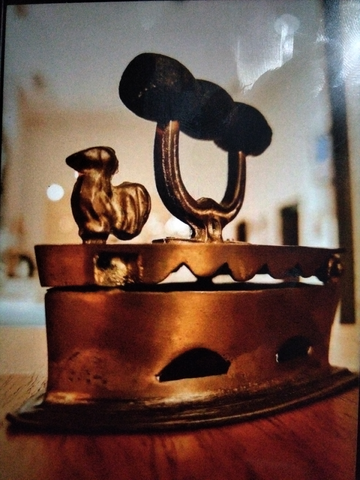 The image shows a close-up of an antique iron with a decorative rooster attached to its top. The iron appears to be made of brass and has a dark handle. The rooster is also metallic and detailed. The iron is placed on a wooden surface, possibly a table. The background is blurred and suggests an indoor setting with soft lighting. There are no people visible in the image. The scene is static and focuses entirely on the object. The provided location in Kualaraja, Indonesia, is not visually verifiable from the image itself.