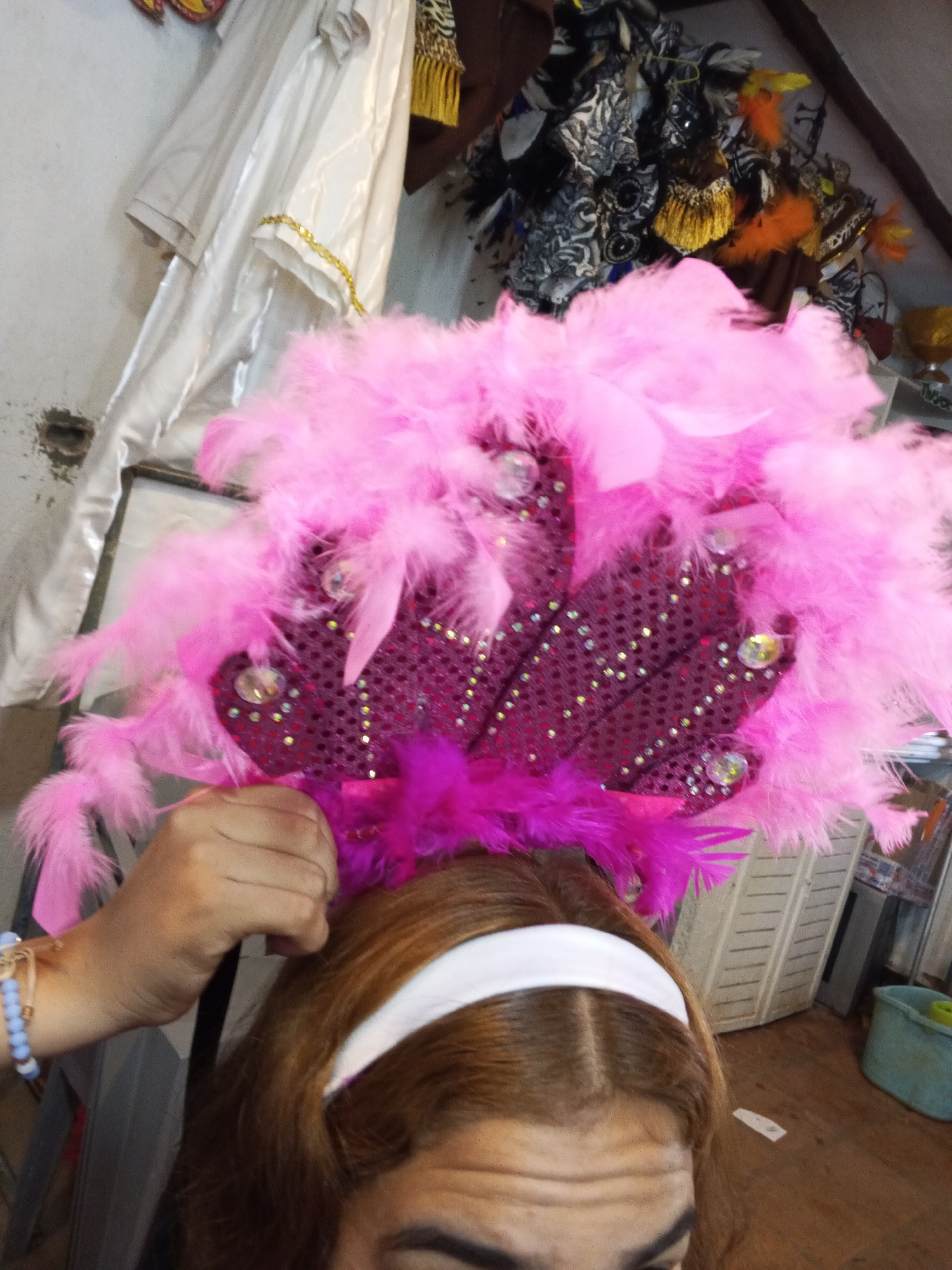 The image features a close-up of a person trying on or adjusting a large, elaborate feathered headpiece, likely in preparation for a festive event in Porlamar, Venezuela. The headpiece is the central focus, characterized by voluminous, bright pink feathers. Its base is a magenta-sequined fabric, embellished with numerous smaller, possibly iridescent gold or yellow sequins, and several larger, clear, faceted gems. Darker pink or purple feathers are visible beneath the main pink plumage, closer to the wearer's head.
A hand, adorned with a light blue beaded bracelet, is visible on the left, holding the headpiece in position. The person's face is partially visible from the forehead upwards, showing brown hair parted in the middle and a white headband. Dark eyebrows are also discernible.
The background suggests an indoor setting, possibly a costume workshop or storage area. On the left, light-colored garments, one with a gold trim, hang against a rough, light-toned wall that has a dark mark. To the right, other decorative items and fabrics, including some with black and white patterns and vibrant orange and yellow feathered or tasseled accents, are partially visible. Further back, a light-colored, slatted storage container and a blue bucket sit on a wooden floor. No discernible text is visible in the image.