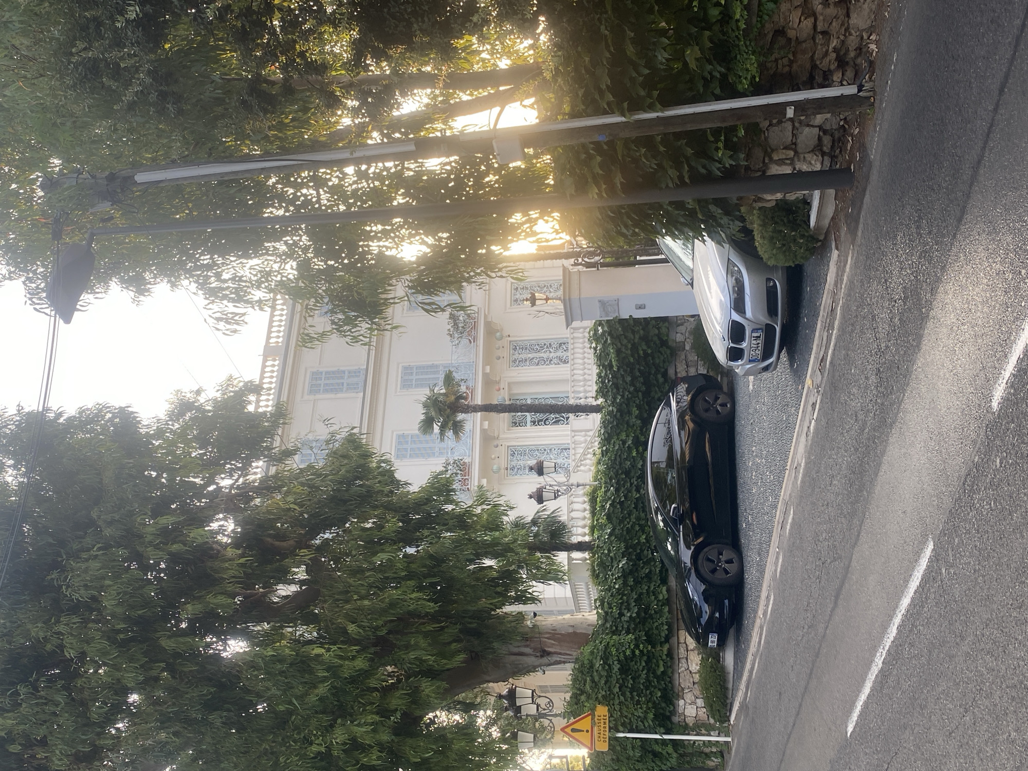 The image depicts a residential street in Antibes, France, likely during daytime as indicated by the sunlight filtering through the trees. A large, white, multi-story building with blue shutters and ornate balconies dominates the background. In front of the building, a stone wall covered in ivy separates it from the street. Two cars are parked on the side of the street: a black Tesla and a silver BMW. The street itself is paved, with a white line marking the lane. Several large trees with dense foliage are present, some of which are obscuring parts of the building and the sky. A street lamp and a yellow traffic sign with a warning symbol and French text indicating "CHAUSSEE DEFORMEE" (deformed road) are also visible on the left side of the image. The scene suggests a quiet, upscale residential area.