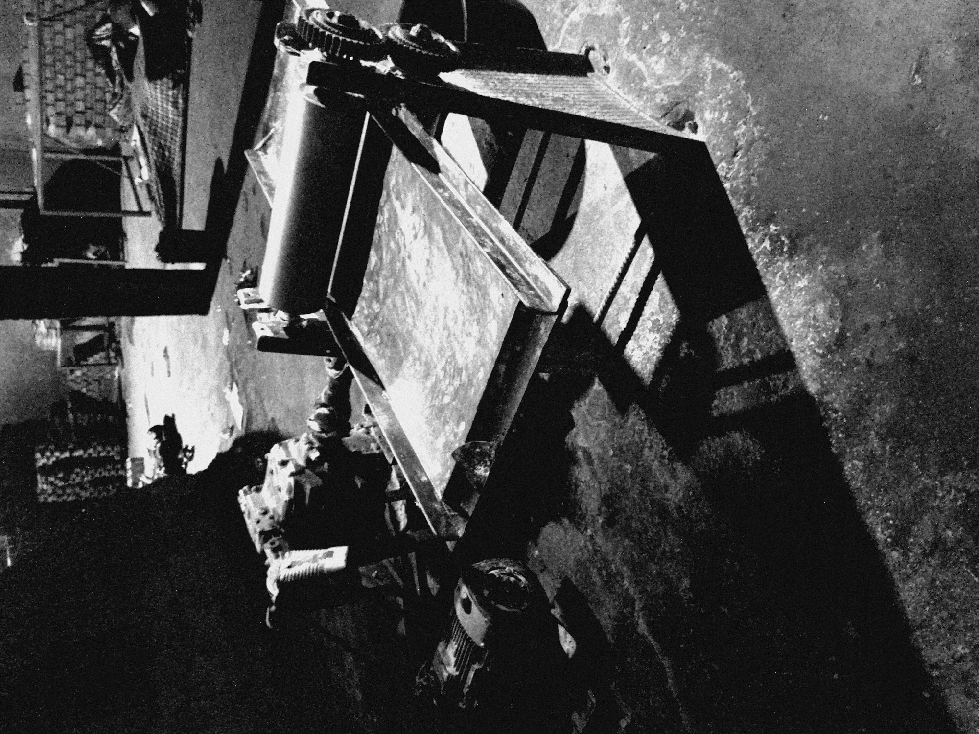 This black and white photograph depicts a stark industrial or workshop setting, characterized by high contrast and noticeable photographic grain. The image primarily features a piece of heavy machinery, likely a press, roller, or grinder, positioned in the midground.
The machine dominates the composition, showcasing large, prominent gears at its upper section, a substantial cylindrical roller below them, and a flat, rectangular metal platform or tray extending outwards. A chain or belt mechanism runs along one side of the machinery, hinting at its operational function. Closer to the foreground, beneath the main structure, a smaller motor or pump unit is visible, connected to the larger apparatus.
Strong, elongated shadows are cast across the rough, possibly concrete or paved floor by the machinery, suggesting a harsh, directed light source originating from outside the frame. These shadows contribute to the dramatic and somewhat gritty atmosphere of the scene.
In the background, which is less defined and slightly out of focus, other elements of an industrial environment can be discerned. These include what appear to be shelving units or racks holding indistinct objects, possibly supplies or equipment parts. A patterned surface, resembling a grate or mesh, is also visible on the left side of the background. The floor exhibits textural variations, indicating wear, dirt, or possibly remnants of liquids.
No individuals are present in the image, and no specific activity is actively taking place; the machinery appears to be idle. The artificial and dramatic lighting, coupled with the black and white rendering, creates a sense of quiet solitude within this functional space. There is no discernible text within the frame.