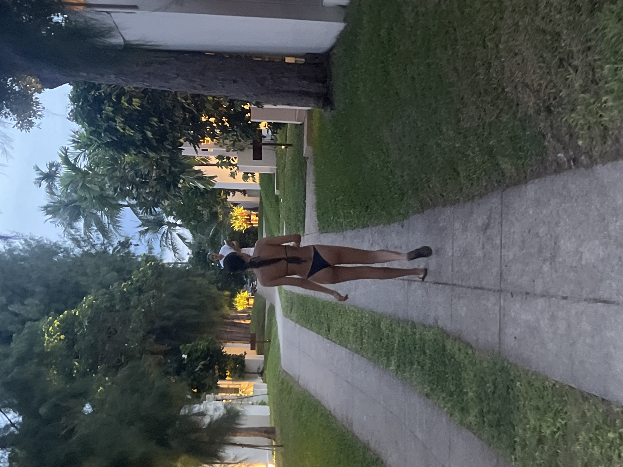 A rear view of a woman in a black bikini walks away from the camera down a concrete pathway bordered by grass. She is wearing flip-flops. In the background, another person in a white shirt stands near some palm trees and foliage. The pathway is in a courtyard or resort area, with white buildings visible on the right side of the image, and trees and vegetation lining both sides of the pathway. The lighting suggests it is dusk or early morning. The location is the district of Ko Samui, Thailand.