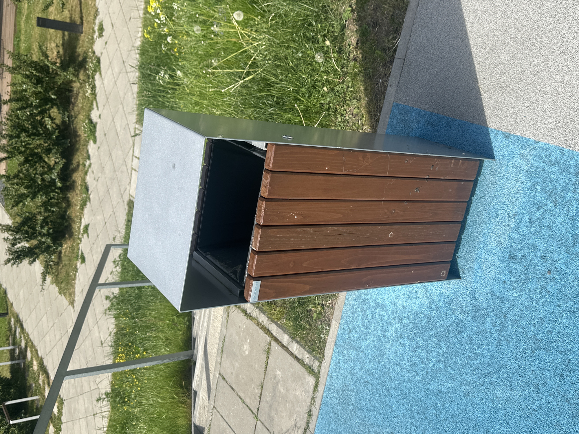 The image shows a modern outdoor trash can with a metal frame and wooden slats on its sides. The trash can is positioned on a blue rubberized surface, adjacent to a gray paved area. In the background, there is a metal railing leading up a walkway paved with square concrete tiles. Lush green grass and some yellow wildflowers grow around the paved area and the railing. Behind the grass, there are dense green bushes. The scene appears to be an outdoor public space, possibly a park, a plaza, or a recreational area. There are no people visible in the image. The lighting suggests it is daytime, with shadows indicating sunlight. The presence of a sign with the brand "JUMST" on the trash can suggests it is a manufactured product.