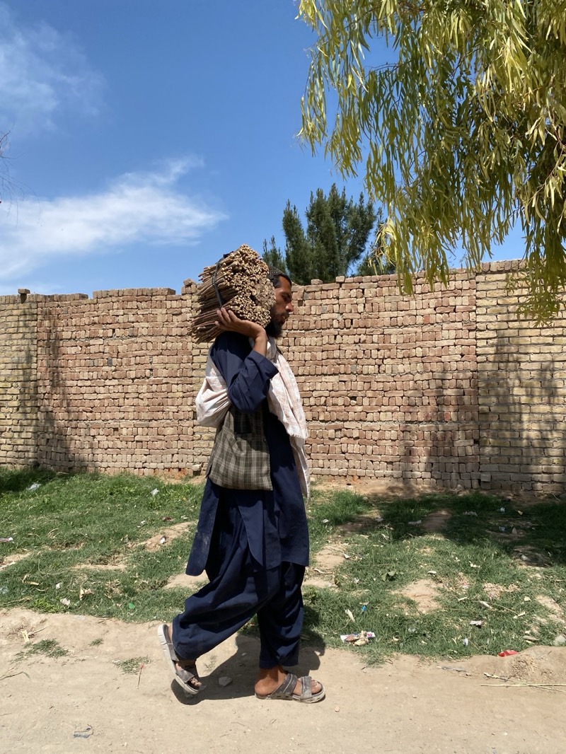 A man walks along a dirt road in Kandahar, Afghanistan. He carries a large bundle of sticks on his left shoulder. He wears a dark blue salwar kameez, a patterned vest, a white scarf, and sandals. A brick wall stands behind him, with grass and trash at its base. A tree's foliage partially obscures the upper right of the frame. The sky is blue with scattered clouds.