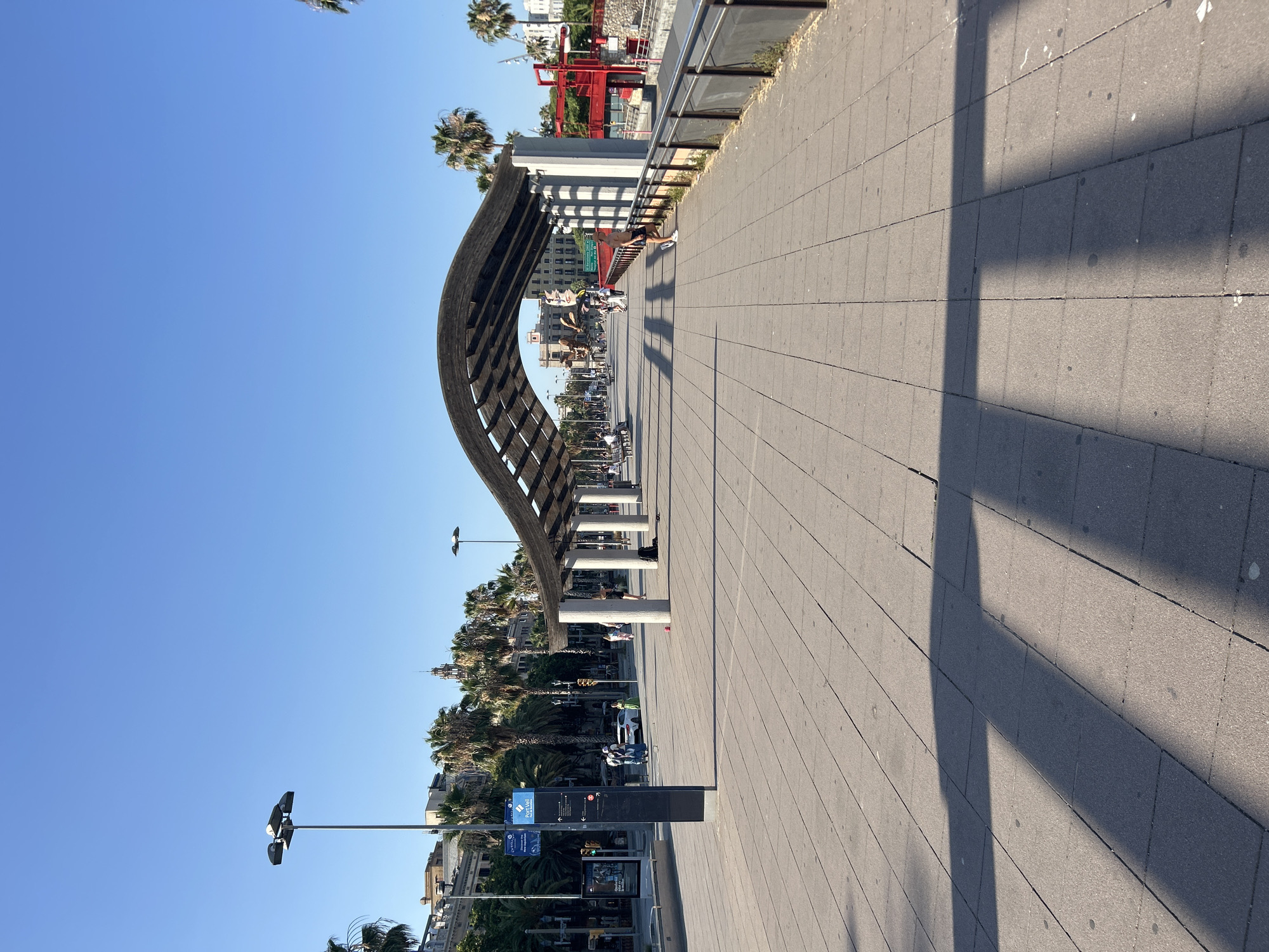 The image captures a sun-drenched promenade in Barcelona, Spain, bathed in the bright light of midday. The scene is dominated by a distinctive wooden structure with a curved, slatted roof, supported by white cylindrical pillars, providing shade along the wide, paved walkway. The walkway, composed of grey rectangular tiles, stretches into the distance, its surface marked by the long shadows cast by the structure and surrounding objects.
Several people are visible on the promenade, engaged in various activities. A man, shirtless and wearing shorts, leans against a railing. Further down, a group of people are seated, while others walk or cycle. The presence of palm trees lining the promenade, along with architectural elements such as buildings with balconies and a tall, spire-like structure in the background, suggest a coastal urban setting.
In the middle ground, there is a street with cars, indicating the promenade is adjacent to a road. A white car is parked, and other vehicles are in motion. On the promenade, there are streetlights with multiple lamps, and signs are visible, including one with the text "Port Vell" and another blue sign with directional arrows and text, likely indicating information about the port.
The sky is a clear, vibrant blue, indicative of fair weather and a sunny day. The overall atmosphere is relaxed and leisurely, characteristic of a pleasant day in a popular public space. The image is oriented vertically, requiring a rotation to view it in its intended landscape format.