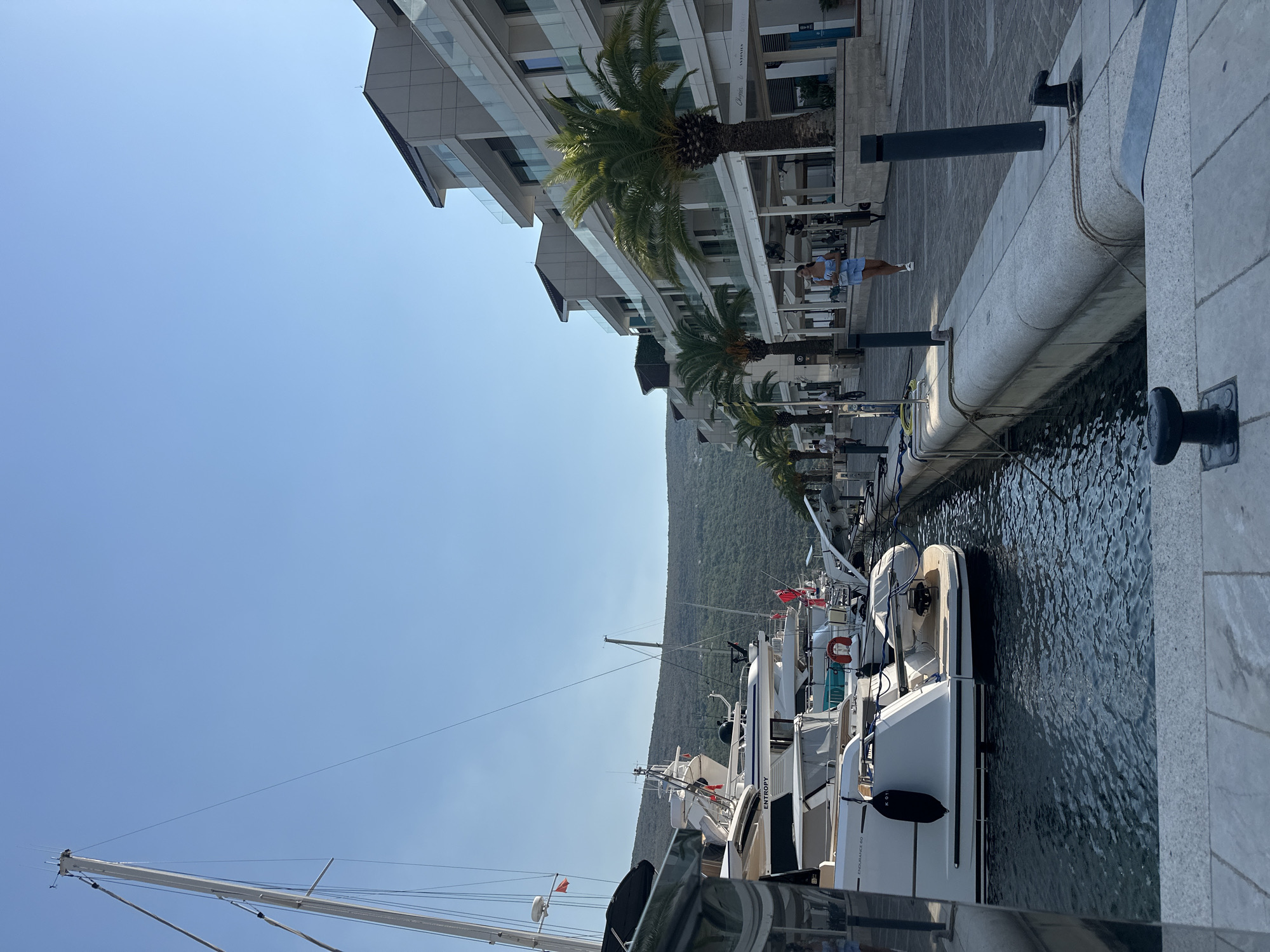 The image shows a waterfront promenade in Kumbor, Montenegro, with a clear blue sky overhead. A row of modern buildings with balconies lines the right side of the scene, interspersed with lush palm trees. In the foreground and middle ground, several yachts and sailboats are docked in the water to the left, their masts and rigging prominent. A paved walkway runs along the waterfront, bordered by a stone seawall. A young woman in a light blue outfit is walking along the promenade, and a few other individuals are visible further down. The overall atmosphere is one of a serene, upscale coastal town.
