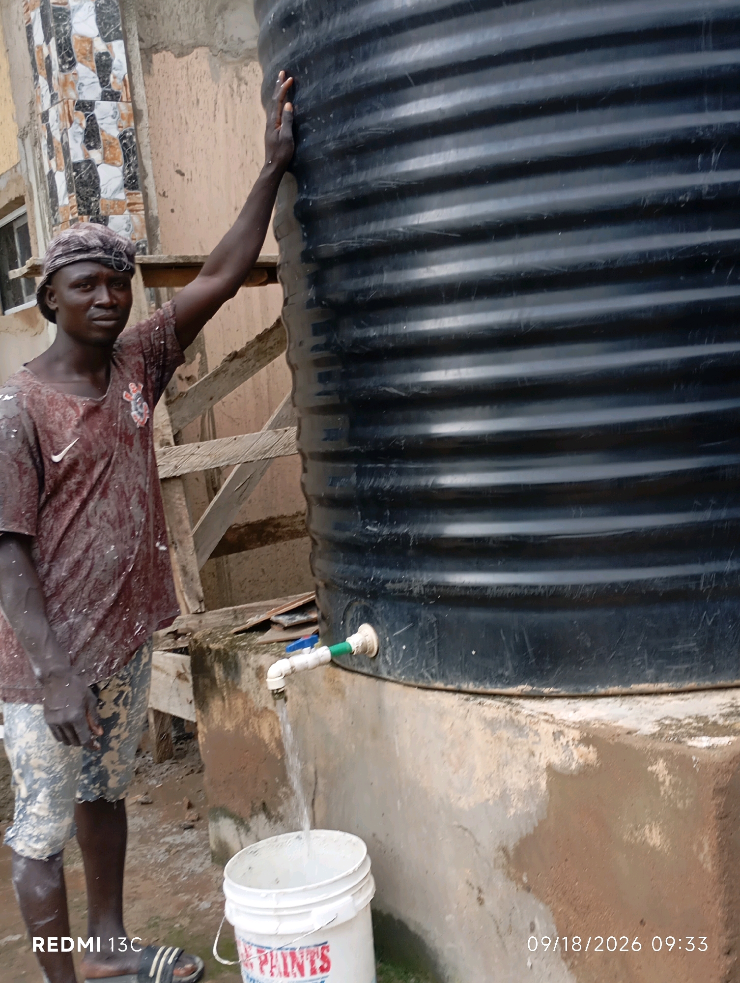 A young Black man wearing a brown, paint-splattered t-shirt and patterned shorts stands next to a large black water tank. He has a light-colored cap on his head and is looking towards the camera with a neutral expression. His left hand is resting on the side of the water tank, and his right arm is bent at the elbow. The water tank is ribbed and appears to be made of plastic. A white tap is attached to the tank, and a stream of water is flowing from it into a white bucket positioned below. The bucket has some lettering on it, partially obscured. The ground around the tap and bucket is wet and muddy. In the background, there is a building with tiled walls and wooden scaffolding. The scene appears to be outdoors, possibly in a residential area or construction site. The timestamp on the image indicates it was taken on September 18, 2026, at 09:33 AM.