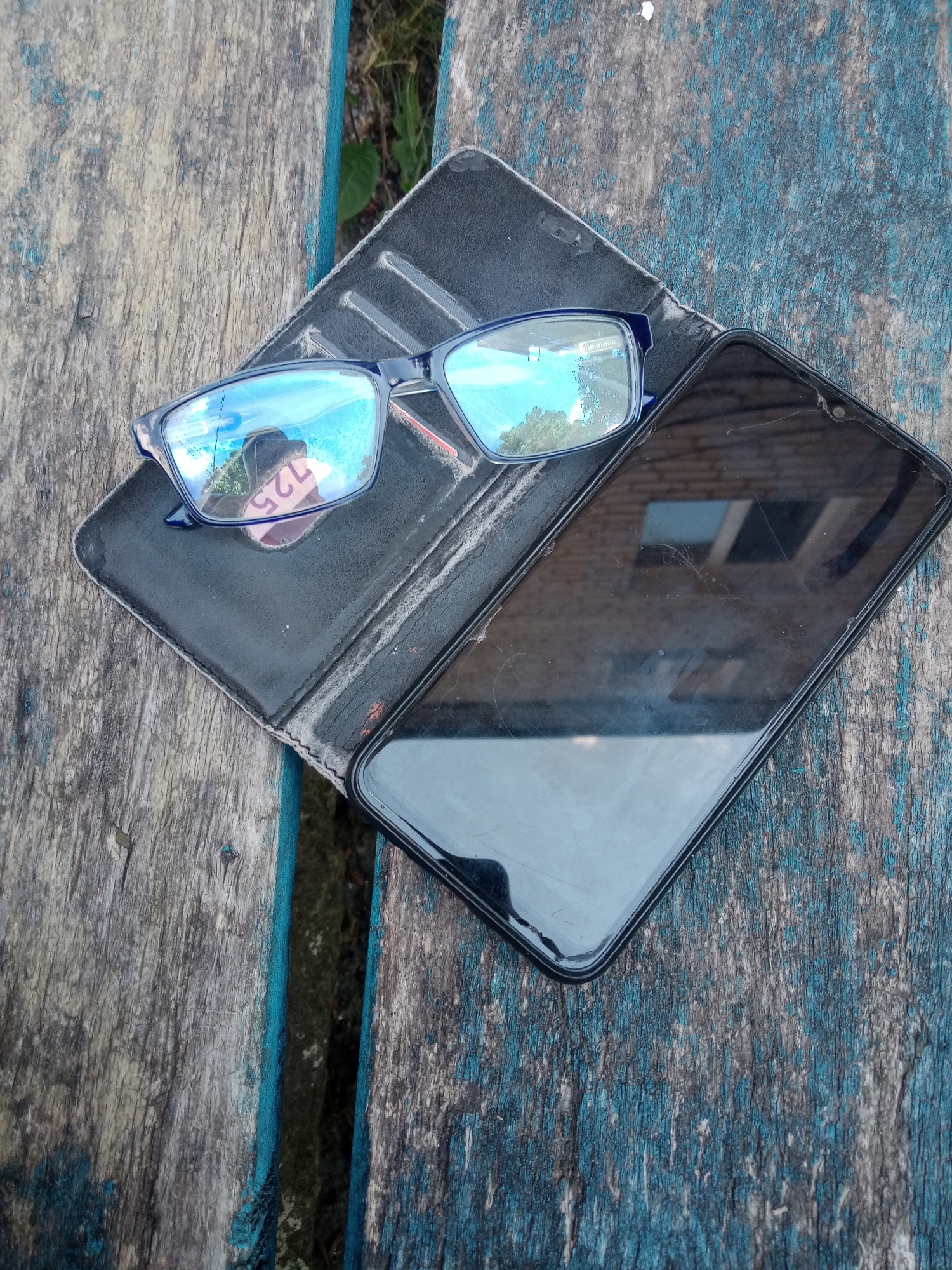 A pair of blue-rimmed eyeglasses and a smartphone are placed on a weathered, blue-painted wooden surface. The eyeglasses are open, with their lenses reflecting the sky and trees. The smartphone is closed, lying face down with its screen exhibiting minor scratches. The wooden surface appears to be part of an outdoor bench or table, with visible grain and chipped paint. The background shows glimpses of foliage and hints of the urban environment of Vlasivka, Ukraine.
