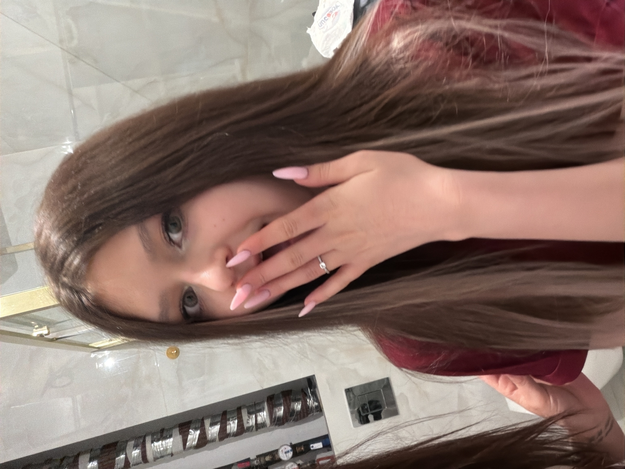 A young woman with long brown hair, green eyes, and pink stiletto nails is posing for a selfie. She is wearing a ring on her finger and is partially covering her face with her hand. The background features a white tile wall, a shelf with various objects, and a light fixture. Based on the objects and architectural style, it is possible this image was taken indoors in Moscow, Russia.