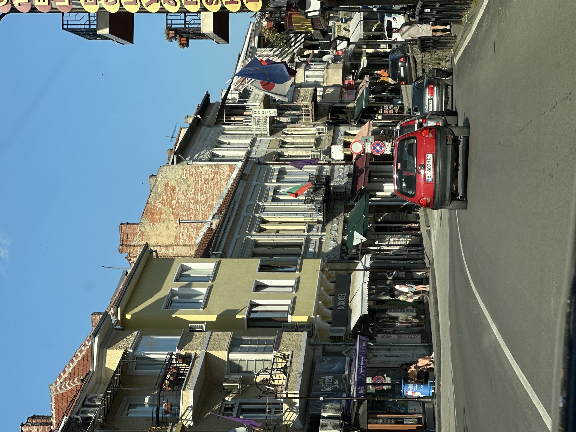 This image captures a street scene in Veliko Tarnovo, Bulgaria, as indicated by the "BT" prefix on the license plate "BT 8024 BT" on the red vehicle in the foreground. The perspective is from inside a vehicle, looking down a street that gently curves to the right. The day is clear and sunny with a bright blue sky, revealing architectural details and casting distinct shadows. The street is lined with multi-story buildings, predominantly commercial on the ground floor with residential or mixed-use above. On the left side of the street, multiple awnings in various colors (dark blue, red, white) extend over shop entrances. Shop signs visible include "Refresh Brass!" and "KOKODEL". The buildings display diverse facades, ranging from textured yellow to light green and grey, some featuring traditional red-tiled roofs with exposed brick chimneys. Numerous air conditioning units are affixed to the exterior walls. Several balconies with black metal railings are present, some adorned with potted plants. Multiple flags are displayed from these buildings, including a Bulgarian flag (white, green, red horizontal stripes), an EU flag (blue with yellow stars), a Japanese flag (white with a red circle), and a plain purple flag. A vertical sign for "Hostel" with a bed icon and "20 m" is also visible. Street furniture on the left includes a tall, ornate black streetlamp and a low blue bin/bench structure. Two red circular traffic signs are present: one with a white horizontal bar (no entry/closed to vehicles) and another with a blue cross (no stopping). On the right side of the street, a large vertical sign with yellow letters spelling "HOTEL YAN..." is partially visible. This building also features balconies with black metal railings and potted plants. Further down the street, buildings with satellite dishes are apparent. A narrow pavement runs alongside the road, separated from a lower level by a stone wall with a black metal railing. Four individuals are visible on the left pavement. Two are seated on the blue bin/bench: one wearing a light-colored dress and shorts, the other a long dress. Two others stand near a shop entrance, appearing to converse; one wears a white t-shirt and light shorts, the other a patterned shirt and light shorts. On the right pavement, a male individual in a light-colored t-shirt and shorts walks away from the camera. Vehicles populate the street. In the immediate foreground is a red two-door hatchback, likely an Opel