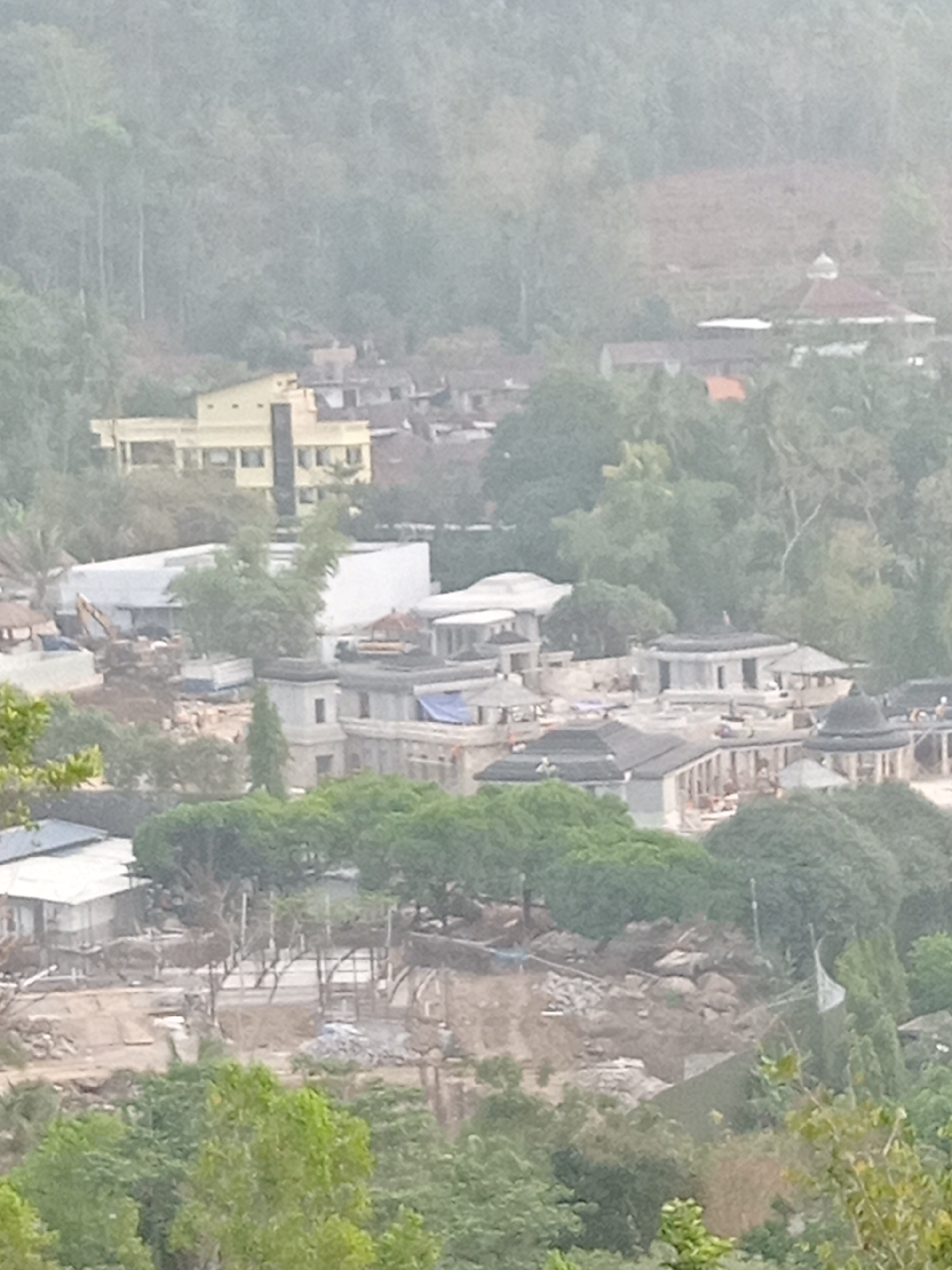 This is a wide-angle, aerial photograph of a developing construction site located in Majaksingi, Indonesia. The scene depicts a collection of buildings in various stages of completion, interspersed with lush green foliage and hilly terrain. In the mid-ground, a prominent light-yellow, multi-story building with visible windows and a dark vertical element stands out. Adjacent to it, several structures appear to be under construction, with exposed frameworks, blue tarpaulins, and piles of building materials. An excavator is visible on the left side of this area. Further into the background, a building with a domed roof, possibly a mosque or religious structure, is partially obscured by trees. The overall environment is a blend of natural landscape and urban development, characteristic of a community undergoing expansion. The image is slightly hazy, suggesting it was taken on a day with limited visibility. No individuals are clearly discernible in the photograph.
