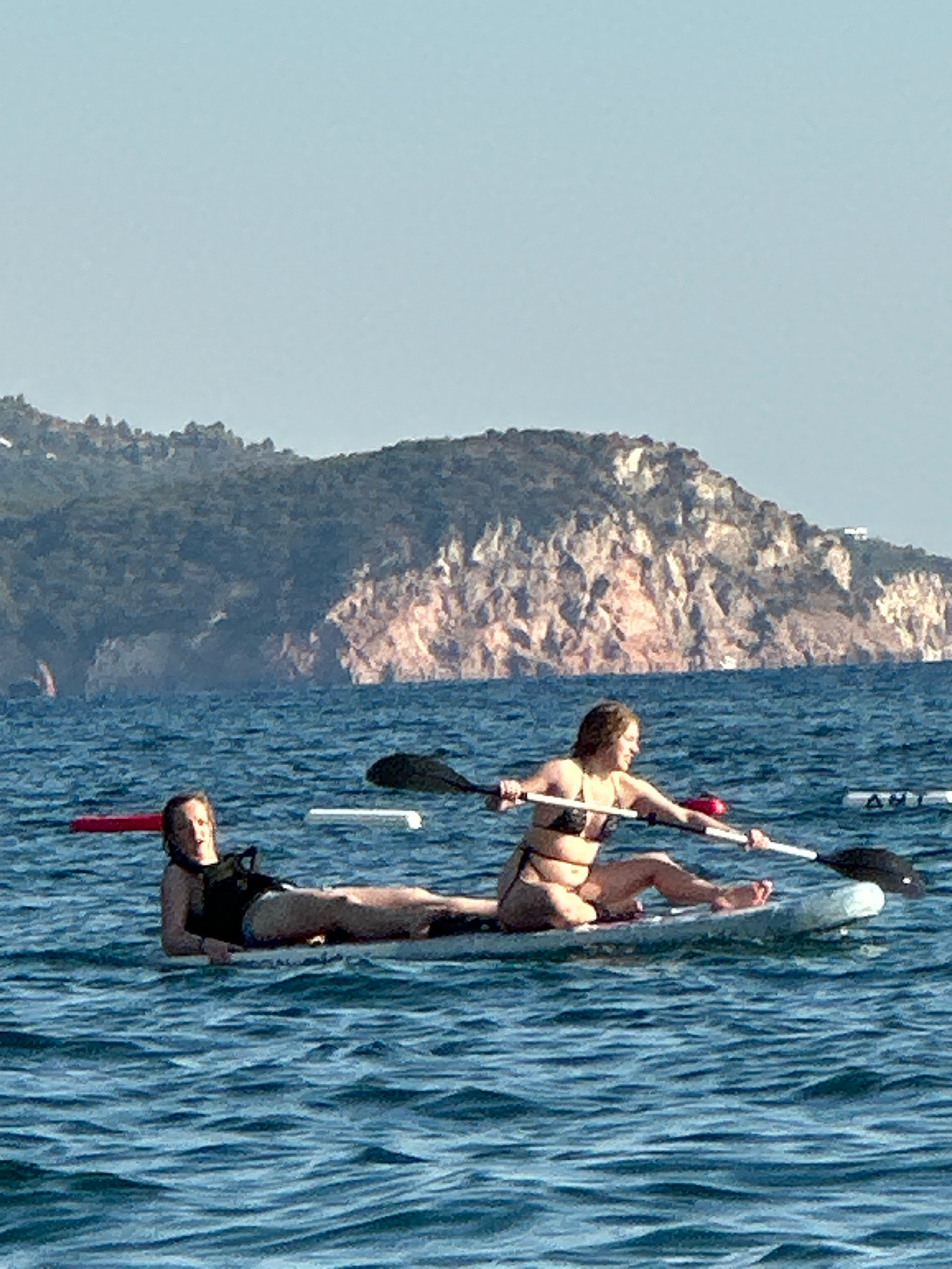 Two women are paddleboarding on the water off the coast of Boreti, Montenegro. The woman in the foreground is wearing a black bikini and is sitting on the paddleboard, paddling with a black and white paddle. The woman behind her is lying on her stomach on the paddleboard, wearing a black swimsuit. The water is blue and choppy, and there is a rocky, tree-covered hill in the background. The sky is clear and blue.