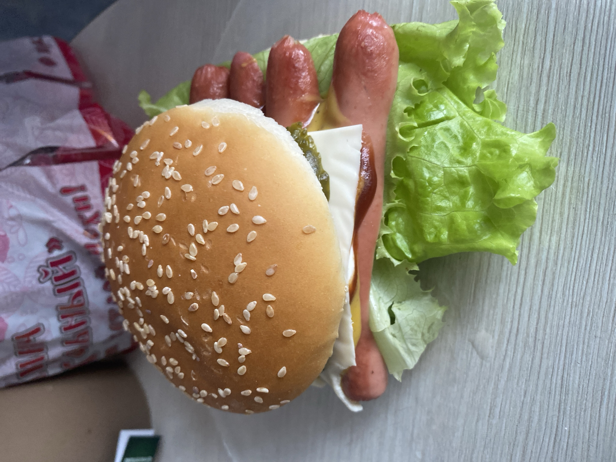The image shows a hot dog placed inside a sesame seed bun, resembling a burger. The hot dog is accompanied by lettuce, a slice of cheese, and what appears to be pickle relish. The setting appears to be indoors, possibly a table or countertop. A fast-food bag with Russian text is partially visible in the background. This image might be associated with a fast food establishment. There is no indication of the location being Izhevsk, Russia.