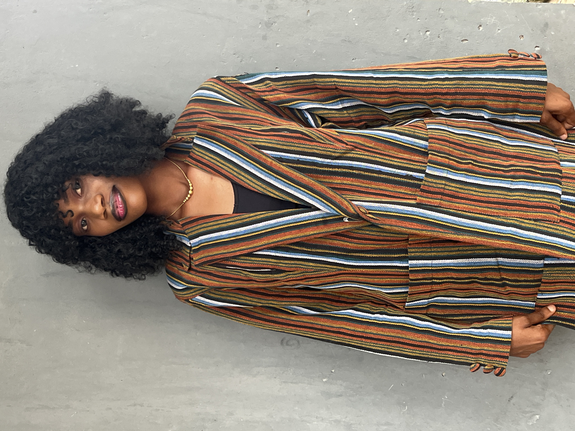 A young Black woman is lying on her side on a grey surface. She has curly black hair and is wearing a striped blazer suit with horizontal stripes of various colors including orange, brown, black, blue, yellow, and white. She is wearing a necklace. The location is Badagry, Nigeria.
