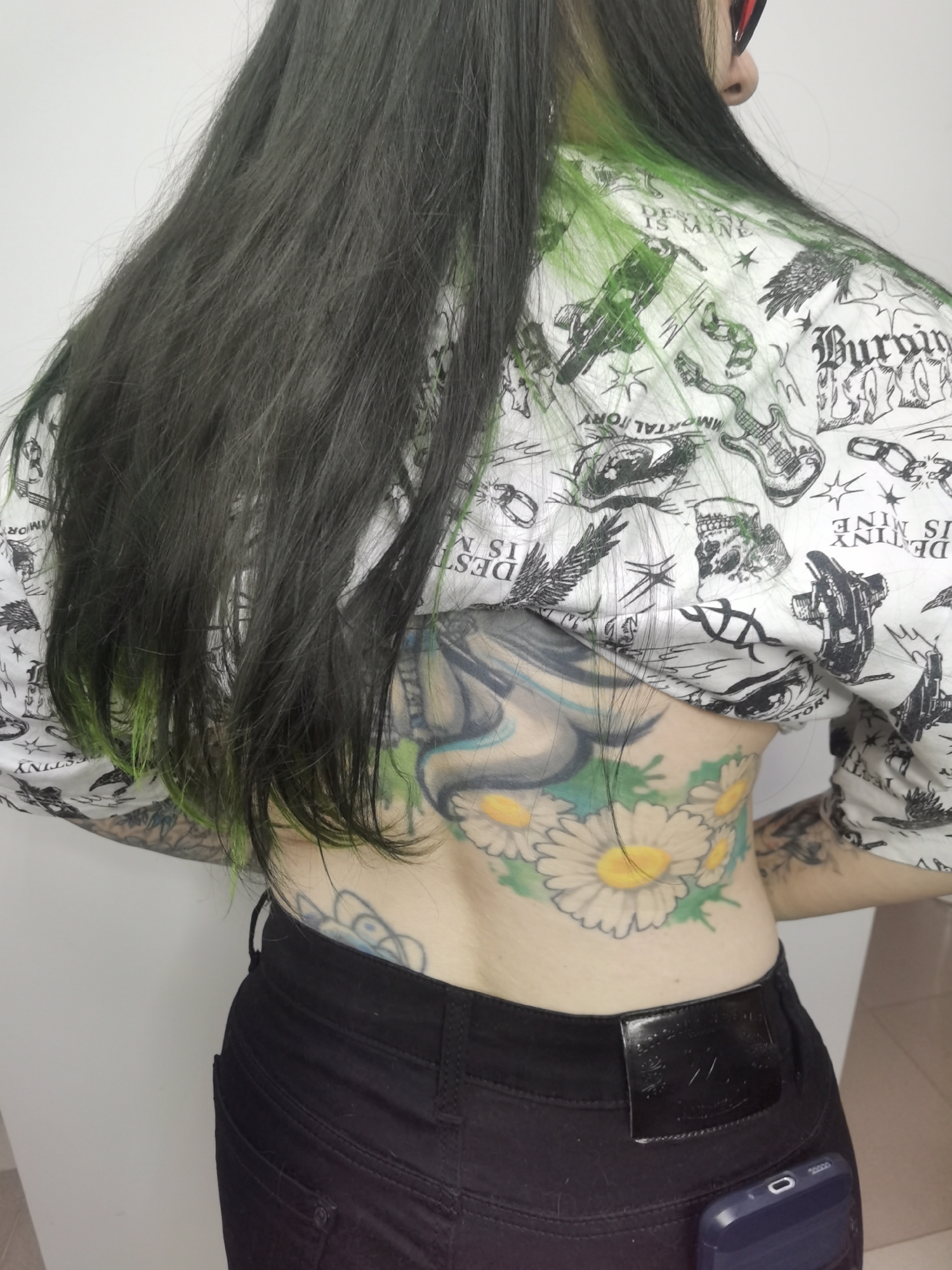 A person stands with their back to the viewer, revealing a tattoo that spans their lower back featuring a large daisy with a yellow center, green leaves, and a blue and white stylized design above it. The individual's hair is long and black, with green highlights near the ends and at the nape of the neck. They are wearing a white shirt with black graphic print and black denim pants. A smartphone in a blue case is visible in the back pocket of the pants. No specific details indicate the location is Chaikovsky, Russia.