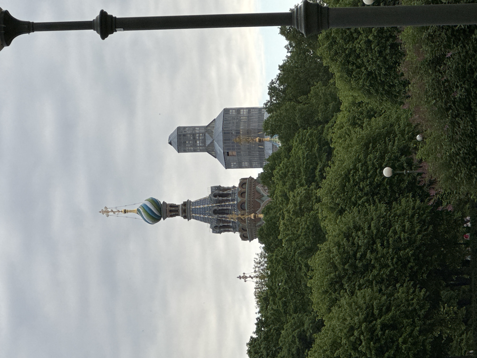 The image, taken in Saint Petersburg, Russia, features two prominent architectural towers rising above a dense canopy of lush green trees under an overcast sky. A dark, ornate lamppost extends across the upper part of the frame.
On the left, a vibrant, multi-domed structure, characteristic of Russian Orthodox architecture, stands out. Its most prominent feature is a large, colorful onion dome, striped in shades of blue and green, topped with an intricate golden finial and cross. Below the dome, the building exhibits rich, detailed ornamentation, possibly mosaics or patterned brickwork, in blues, browns, and greens, with arched windows and decorative elements.
To the right of this ornate building, a second, taller tower is visible. This tower appears to be undergoing renovation or construction, as it is largely encased in a grey, semi-transparent protective sheeting or scaffolding. Golden elements are faintly discernible through the covering.
The lower portion of the image is dominated by thick, verdant green trees, which partially obscure the bases of both structures. Among the foliage on the right, two white spherical lamp fixtures are visible. The sky is a uniform, light grey, indicating an overcast daytime. No people or visible text are present in the scene.
