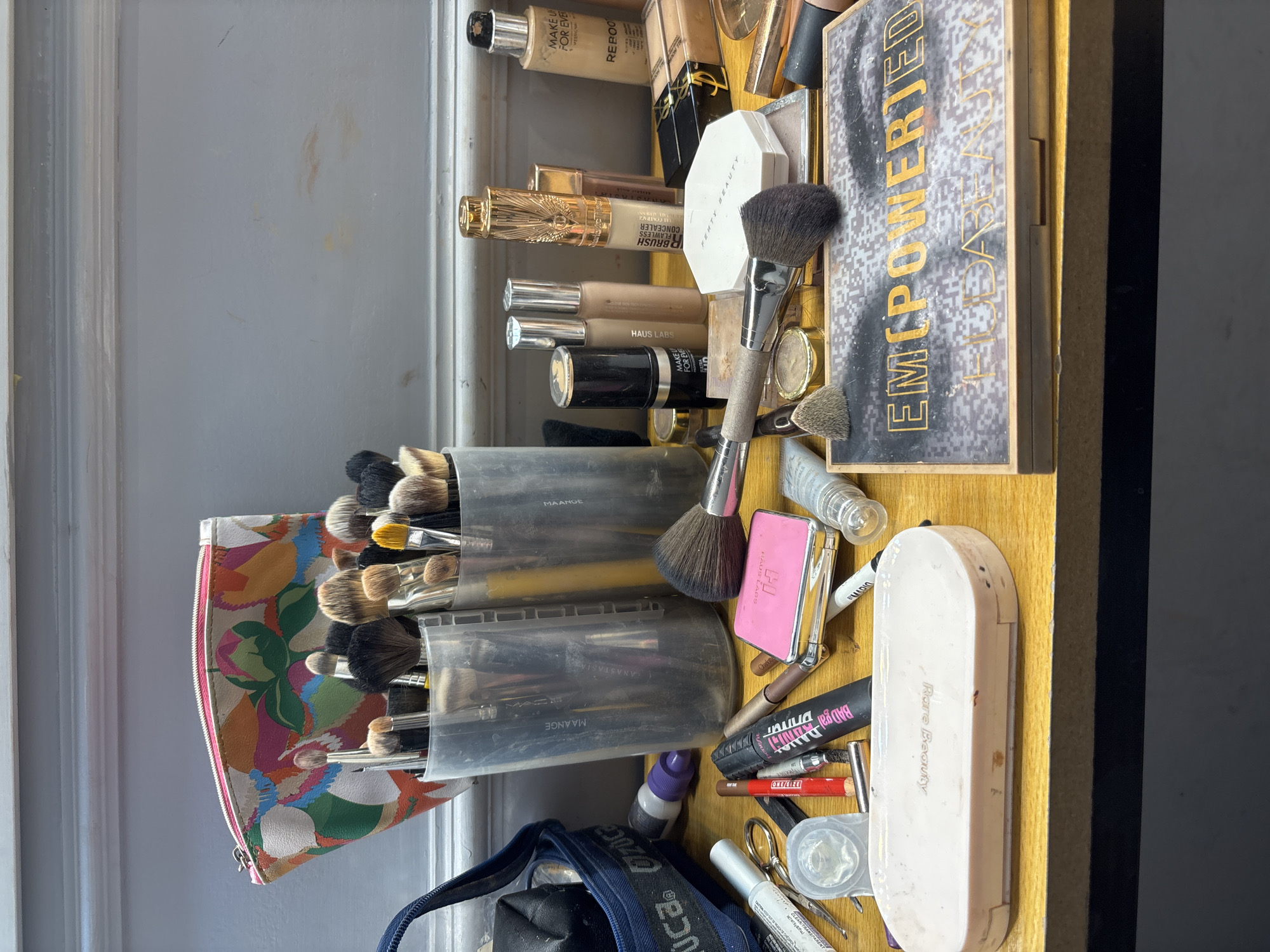 A collection of makeup products is arranged on a wooden surface, likely a vanity or table. In the background, a floral patterned makeup bag sits behind two clear cups filled with makeup brushes. Several makeup bottles and tubes, including foundation and concealer, are scattered across the surface. Also visible are various makeup brushes, an eyeshadow palette, and a compact makeup case. The scene appears to be an indoor setting, with a gray wall in the background. The lighting suggests it is daytime. There are no people present in the image. The location in Sweileh, Jordan, is not discernible from the visual evidence in the image.
