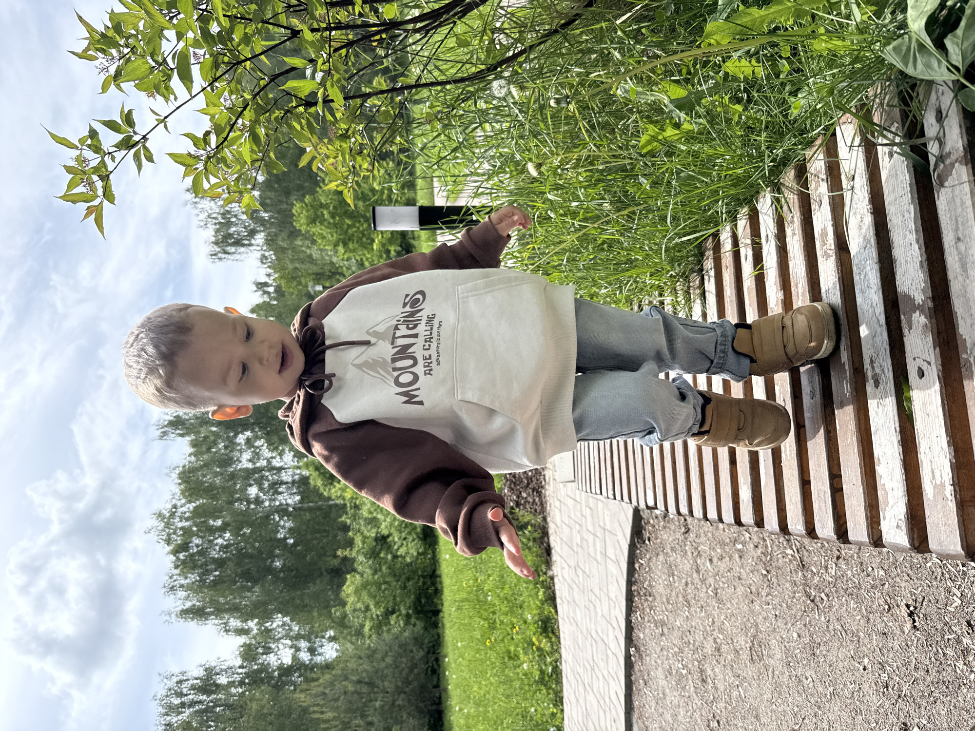 A young child is walking on a wooden bench in a park. The child is wearing a brown and white hoodie with the word "MOUNTAINS" printed on it, blue jeans, and tan shoes. The bench is made of wooden slats and is located next to a paved path. In the background, there is green grass, trees, and a cloudy sky. The photograph appears to be taken in Заволжье, Russia, given the context. A white and black pole is visible in the background.