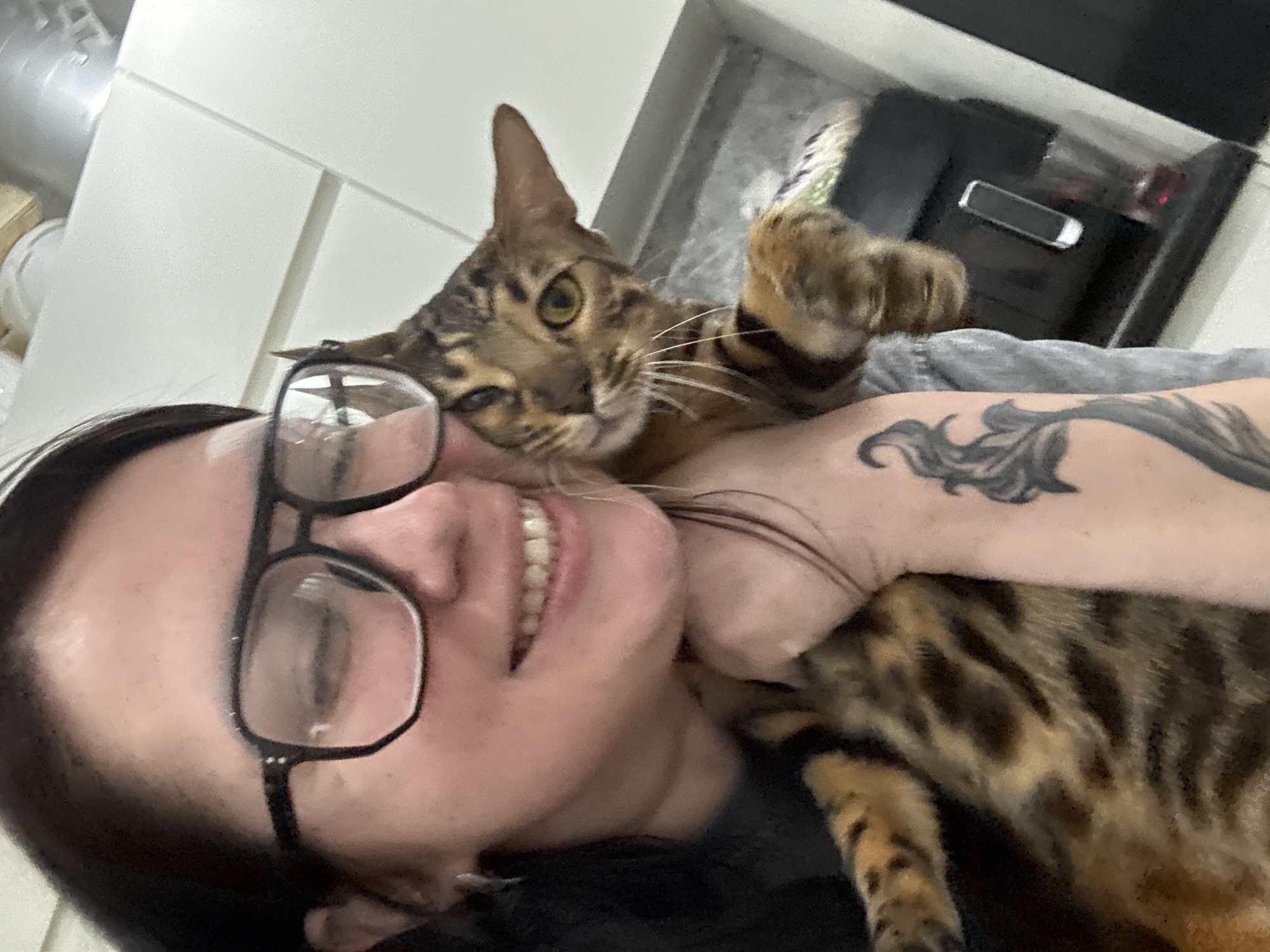 The image is a close-up selfie-style shot of a person and a cat. The person, appearing to be female, is smiling broadly with teeth visible, and wears black-framed glasses. Their dark hair is visible, and a large, dark-toned tattoo with a swirling, organic design is prominent on their right arm.
Cuddling close to the person's face and body is a Bengal cat, identifiable by its distinctive spotted and striped fur pattern in shades of brown and black. The cat has striking light green or amber eyes and is looking towards the camera. One of the cat's front paws is raised and positioned near the person's face or neck, suggesting a playful or affectionate gesture.
The setting is indoors. In the background, on the left, are white rectangular panels or tiles, possibly part of a wall or kitchen cabinetry. On the right, a darker, indistinct area features a small, light-colored object resembling a mobile phone or remote control resting on a dark surface. The lighting is bright and even, highlighting the subjects. The overall emotion conveyed is one of warmth and happiness, stemming from the interaction between the person and their pet. No visible text is present in the image.