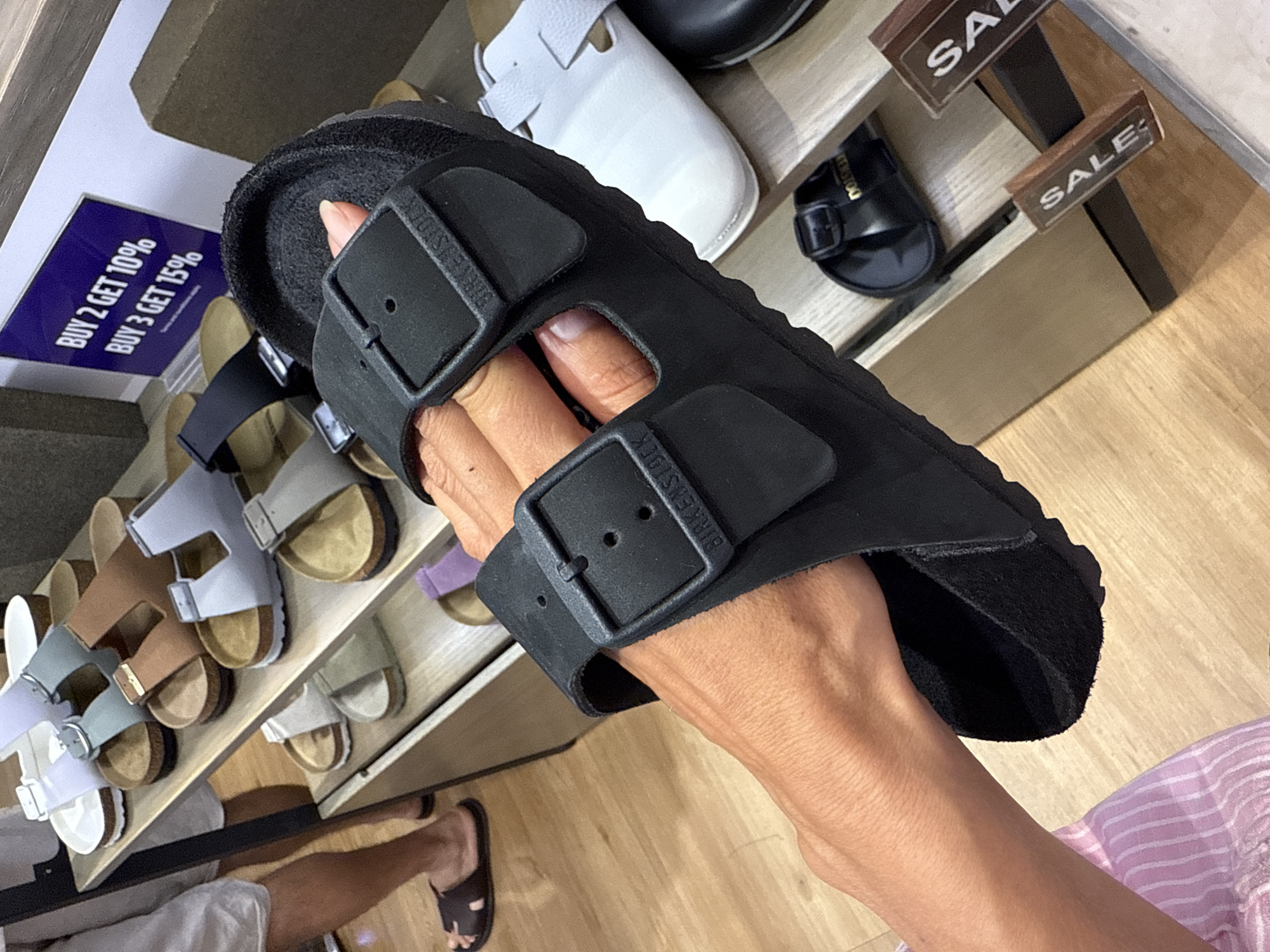 A person's hand is holding up a black Birkenstock sandal against a backdrop of a retail store shelving unit displaying various sandals. The sandal in the foreground features two adjustable black straps with silver buckles, with the "BIRKENSTOCK" logo embossed on the inner strap. The shelving unit contains multiple pairs of sandals in different colors, including white, light blue, and brown, some with cork soles. To the left, a person's legs and feet are visible, one wearing a dark brown sandal. A blue sign in the background advertises a "BUY 2 GET 10% BUY 3 GET 15%" sale. Two wooden "SALE" signs are visible on lower shelves. The environment appears to be indoors, suggesting a shoe store or department store. The location is identified as Nha Trang Ward, Vietnam.