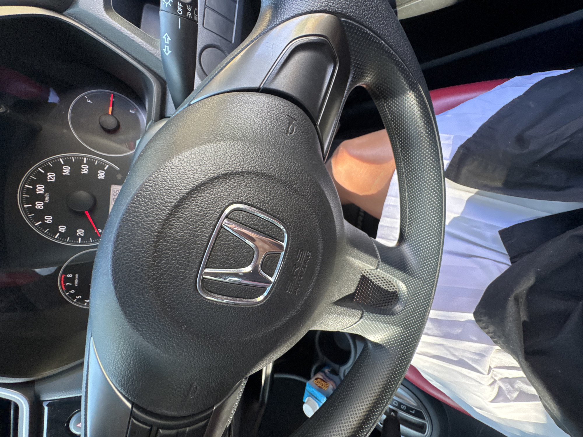 Here is a description of the image:
The image depicts the interior of a car, specifically the steering wheel of a Honda vehicle, with the Honda logo prominently displayed in the center. To the left of the steering wheel, a portion of the dashboard is visible, including the speedometer and fuel gauge. The fuel gauge indicates a low fuel level. The gear shift is visible to the bottom right of the steering wheel. There is also a bottle of Hydro Coco coconut water. Parts of the driver's clothes are also visible on the right side of the image.
Without further information, it is impossible to say whether the picture was taken in Denpasar, Indonesia.
