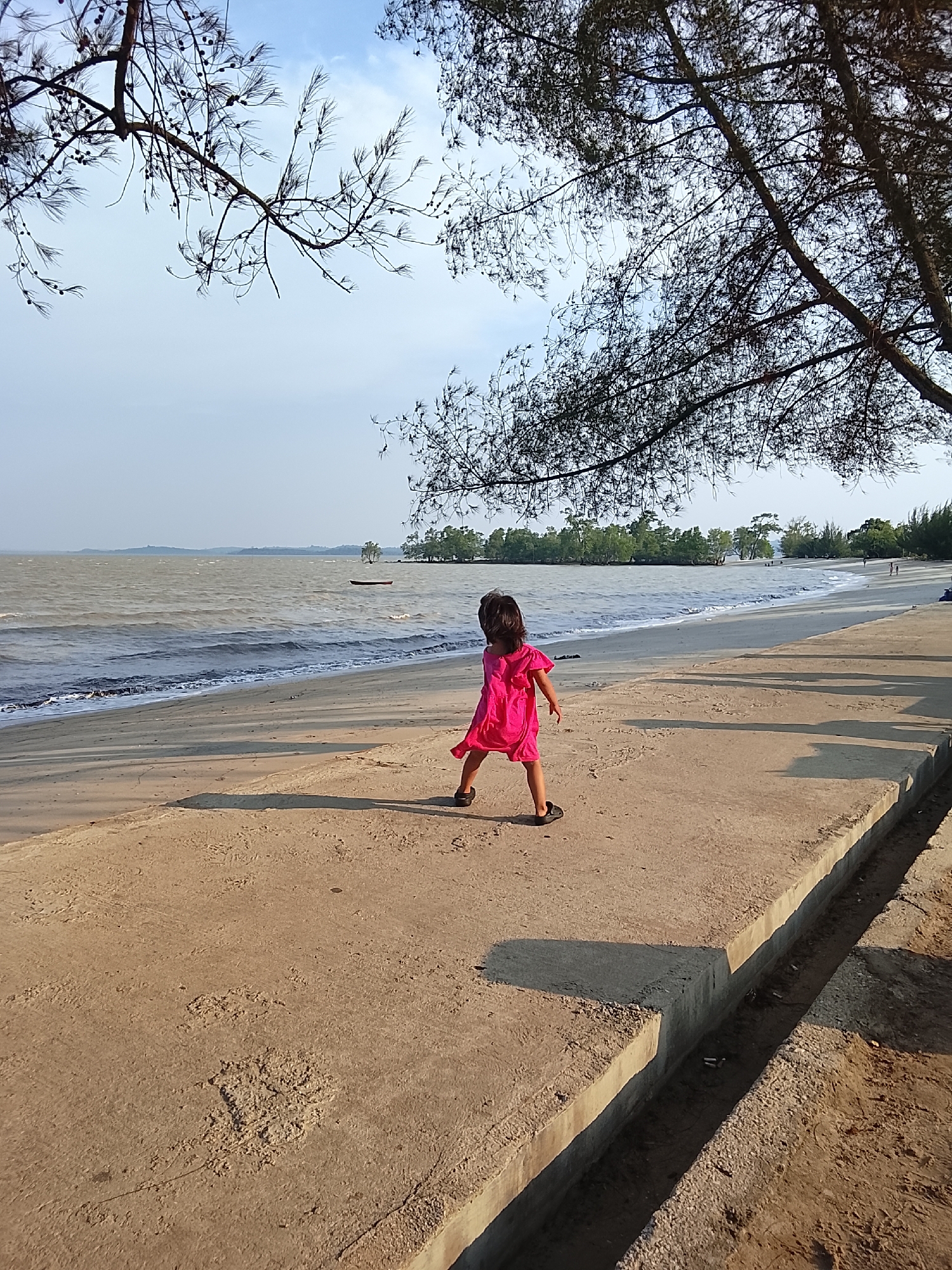 A young girl in a bright pink dress stands on a concrete pathway at the edge of a sandy beach. She is facing away from the camera, looking out at the choppy, gray water of the sea. In the distance, a small boat is visible on the water, and a line of trees separates the sea from the sky. The sky is a pale blue with some wispy clouds. The concrete pathway is rough and textured, with visible debris on its surface. The overall scene suggests a slightly overcast day at a coastal location. The location is the beach in Lubuk Puding, Indonesia.