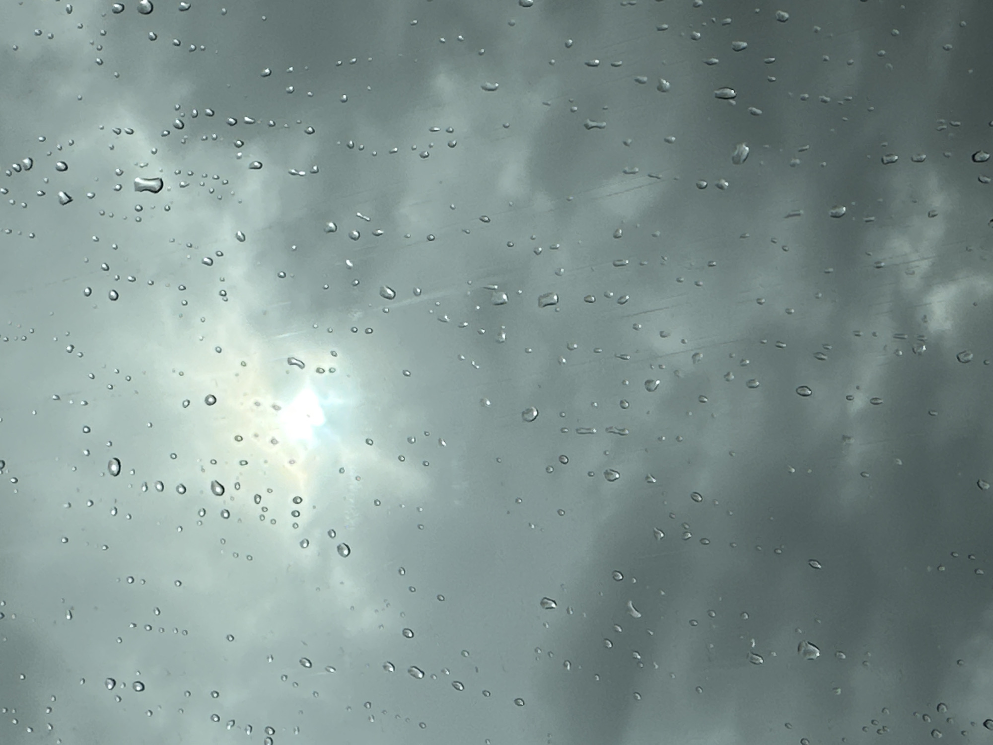 The image shows a close-up view of raindrops on a glass surface, likely a window, with a cloudy sky and a bright sun partially obscured by clouds in the background. The raindrops are scattered across the glass, creating a textured effect. The overall impression is one of a rainy or overcast day. No specific location in Makiv, Ukraine is identifiable from the image.