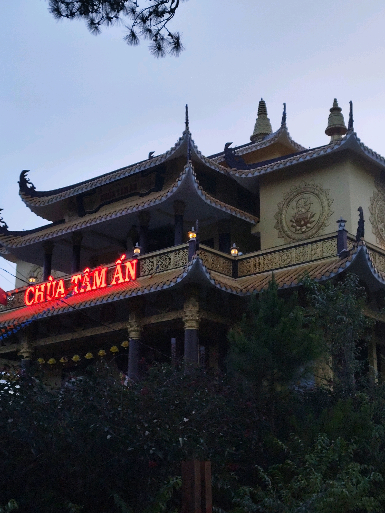 The image shows a large, ornate building with multiple levels and a traditional Asian architectural style. The building is predominantly yellow and gold, with dark blue or black roofs that have intricate decorative elements. Red neon lights spell out "CHUA TAM AN" across the front of one of the upper levels. The building is surrounded by lush green foliage, with trees and bushes visible in the foreground and obscuring parts of the lower levels. The sky is a pale blue-gray, suggesting it may be dusk or early morning. Decorative lanterns are visible on some of the balconies, and elaborate circular motifs are carved into the exterior walls. The overall impression is of a significant religious or cultural structure. This building is Chua Tam An, a temple located in Da Lat, Vietnam.