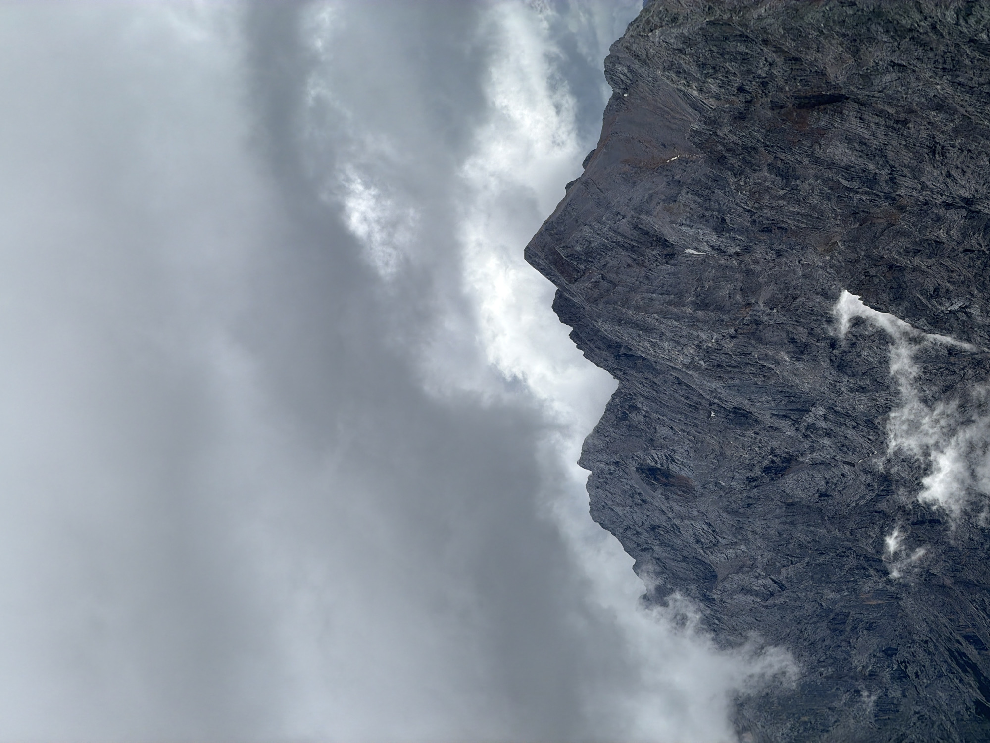 This is a vertical, medium-shot photograph of the jagged, rocky peaks of the Alps under a cloudy sky. The mountains occupy the bottom two-thirds of the frame, with their dark gray, textured surfaces illuminated from above by the overcast sky, creating strong shadows and highlights that emphasize their ruggedness. Wisps of white fog or clouds cling to the lower slopes and crevices of the mountains. The sky above is filled with heavy, dark gray clouds, with some lighter patches suggesting diffused sunlight. The scene appears to be a natural landscape, likely at a high elevation. Given the description of jagged, rocky peaks and a cloudy alpine sky, this image could depict a scene in the Alps near Garmisch-Partenkirchen, Germany, a region known for its mountainous terrain and dramatic weather. There are no people or man-made objects visible in the image.