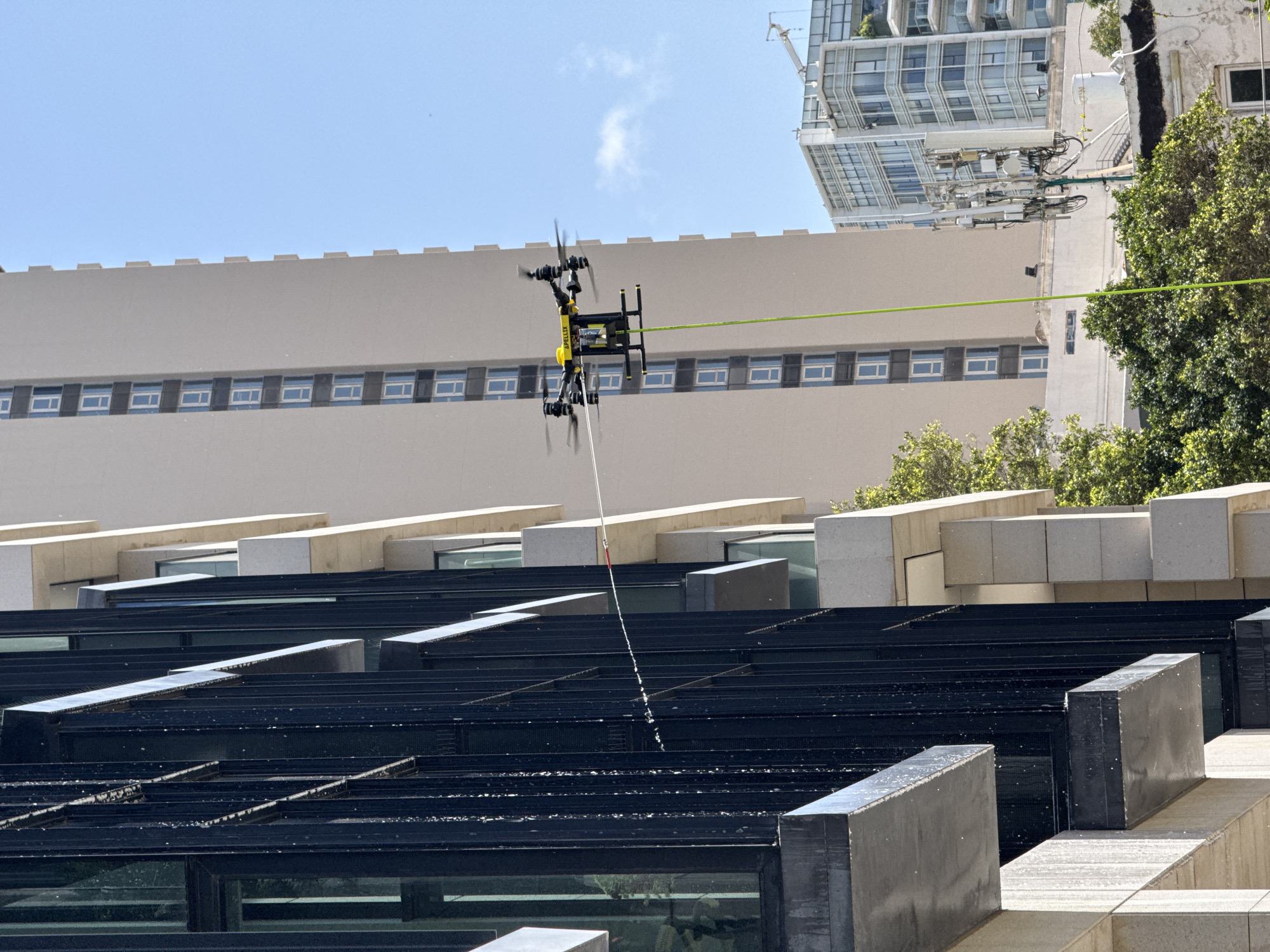 A yellow and black multi-rotor drone, with the word "APELIX" visible on its side, is captured in mid-flight under a clear blue sky with a few wispy clouds. Its propellers are blurred from motion as it hovers. A long, white pole extends vertically downwards from the drone, actively spraying water onto the architectural elements below. A bright green cable or tether extends horizontally from the drone towards the right of the frame, possibly indicating it is tethered for power or guidance.
In the background, a large, beige building with a uniform row of small, square windows spans horizontally. To the upper right, a modern high-rise building with a glass and steel facade is partially visible. An older building section to the right hosts a dense cluster of telecommunications equipment, partially obscured by green foliage.
The foreground and midground are dominated by a dark, multi-layered architectural structure, likely a roof or an elaborate water feature, composed of parallel black panels and interspersed light-colored concrete blocks. Water droplets are visible on these surfaces, indicating the impact of the drone's spray. The scene is brightly lit by natural sunlight, suggesting a clear day in an urban environment, consistent with Tel-Aviv, Israel. The drone appears to be engaged in a cleaning or maintenance operation on the building's exterior or roof.