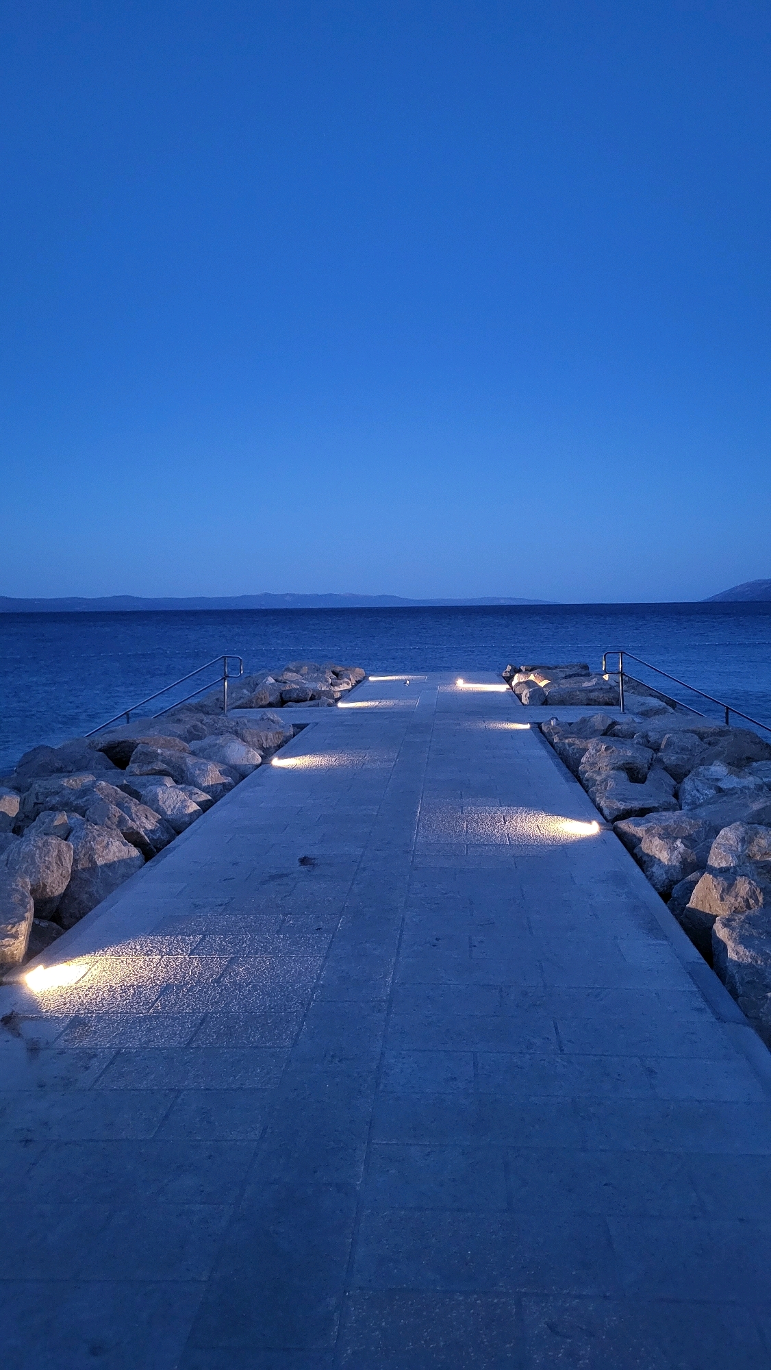 This is a vertical, medium-shot, outdoor photograph taken at dusk in Općina Tučepi, Croatia. The scene depicts a modern paved walkway leading into the calm sea, flanked by large, grey rocks on either side. Built-in lights illuminate sections of the walkway, casting pools of light on the pavement. The sea is a deep blue, meeting a dark blue sky with a faint line of distant hills on the horizon. The walkway appears to be a pier or a promenade constructed on a breakwater. No people or animals are visible in the image. The overall mood is serene and tranquil, highlighting the architecture and natural beauty of the coastal location.